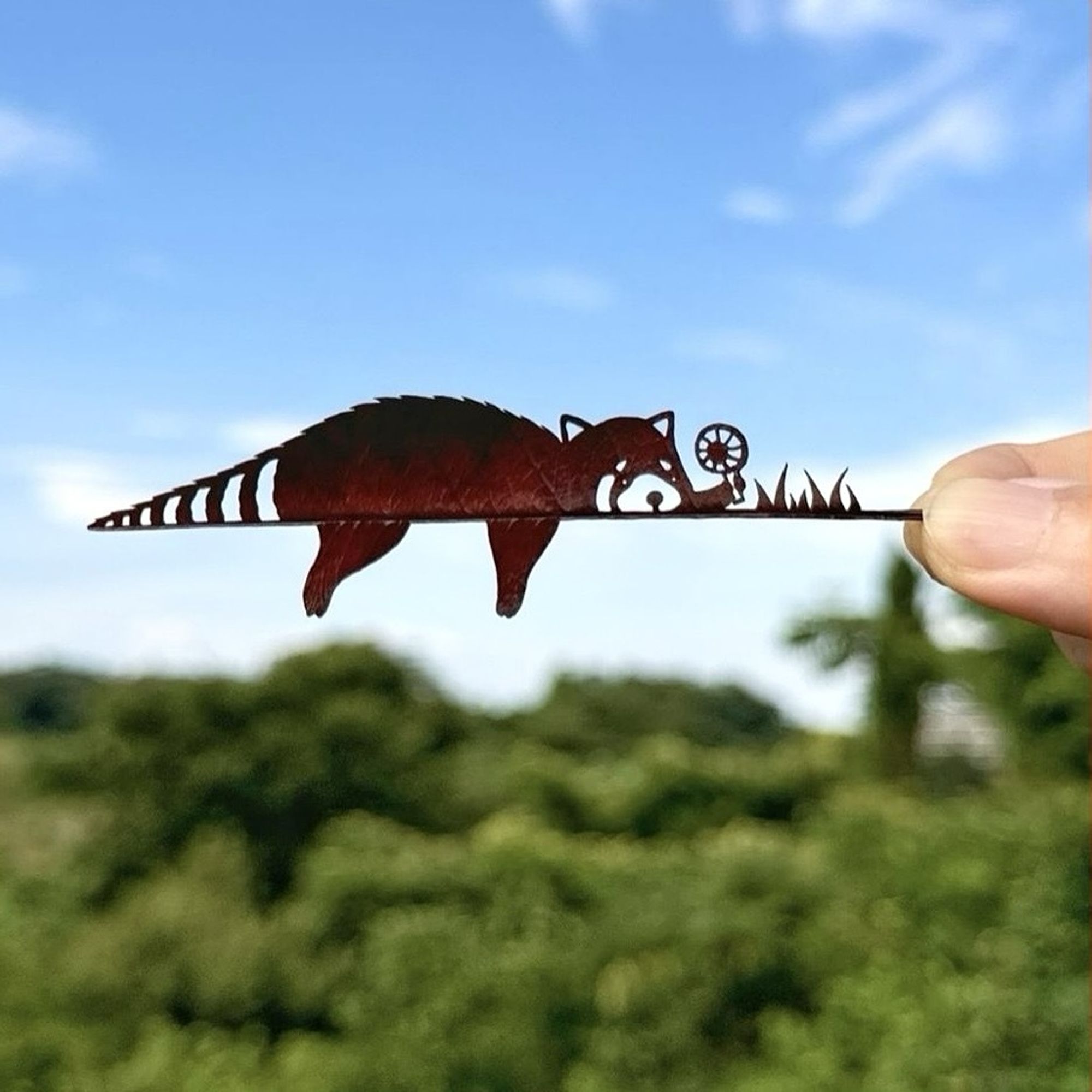 A brown leaf is held horizontally in front of a blue sky.  The top half is a raccoon lying flat on his tummy with his legs hanging off the midrib of the leaf.
He is holding a flower in front of him and there are also a few blades of gras.