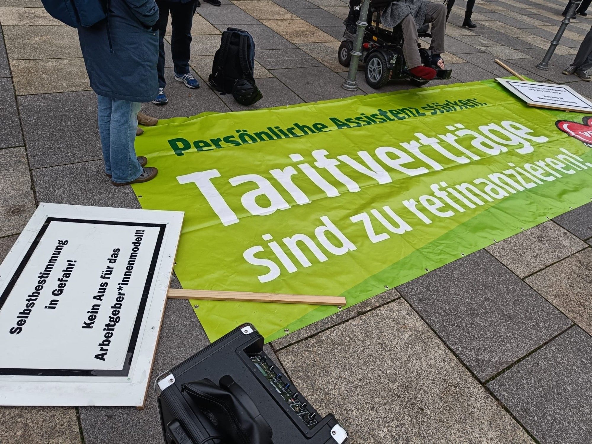 Ein grünes Demotransparent, darauf in dunkel grüner Schrift: Persönliche Assistenz stärken, darunter in großen weißen Buchstaben: Tarifverträge sind zu refinanzieren! daneben ein Demoschild mit der Schrift: Selbstbestimmung in Gefahr! kein Aus für das Arbeitgeber*innenmodell