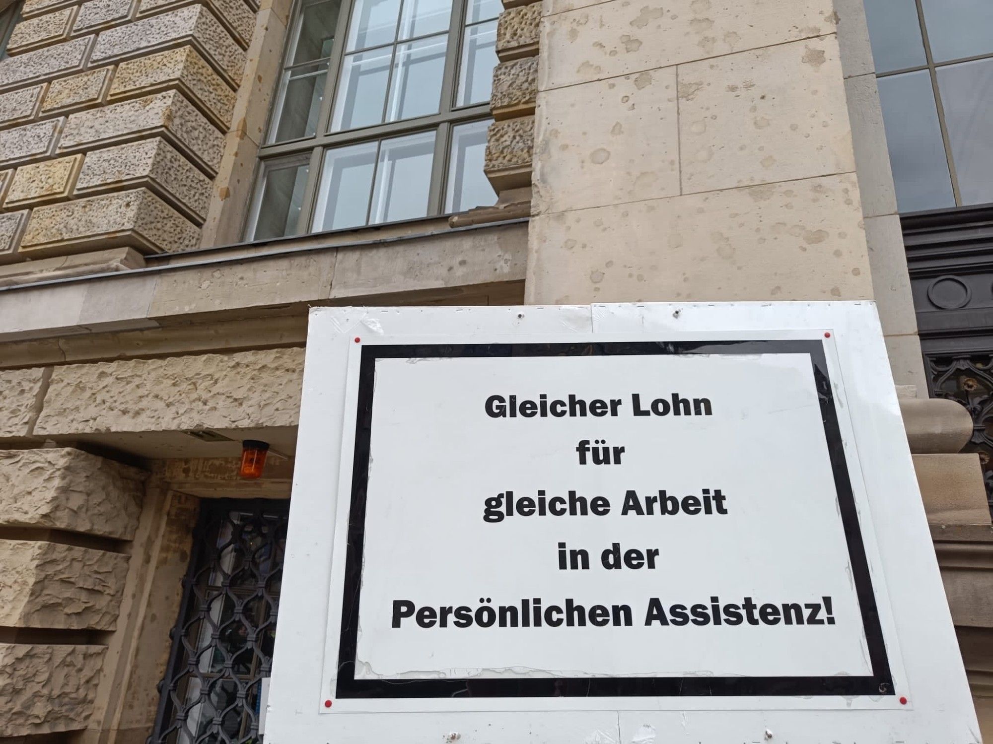 Demoschild vor einem Regierungsgebäude: Gleicher Lohn für gleiche Arbeit in der persönlichen Assistenz!