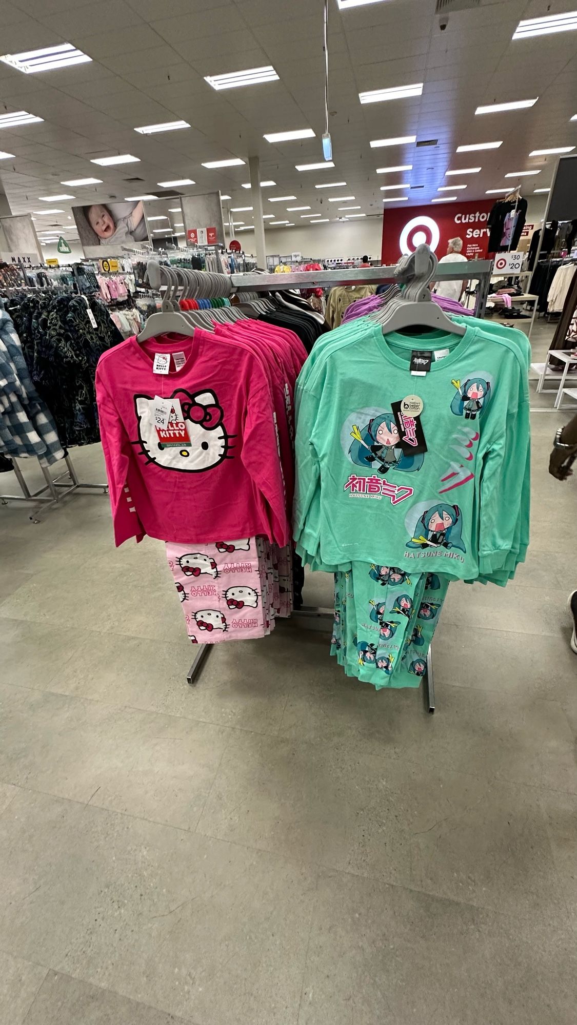 A store clothing rack with two children’s pyjamas being hung on sale. Left side is Hello Kitty sold in pink and right side is Hatsune Miku sold in mint green. Both sets have their respective character designs or derivatives on there.