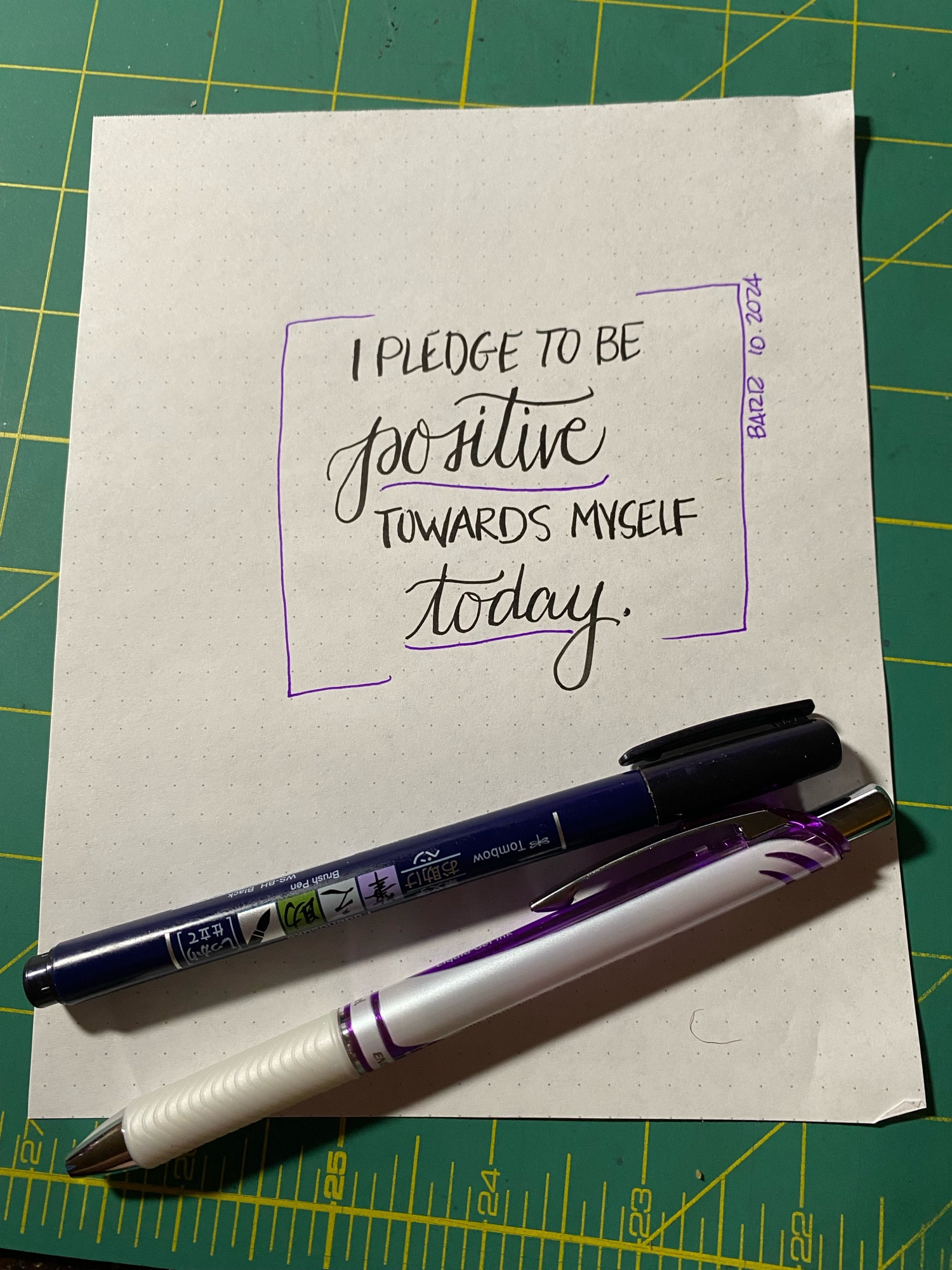 Handwriting practice on a sheet of paper with two pens, text reads “I PLEDGE TO BE POSITIVE TOWARDS MYSELF TODAY.”