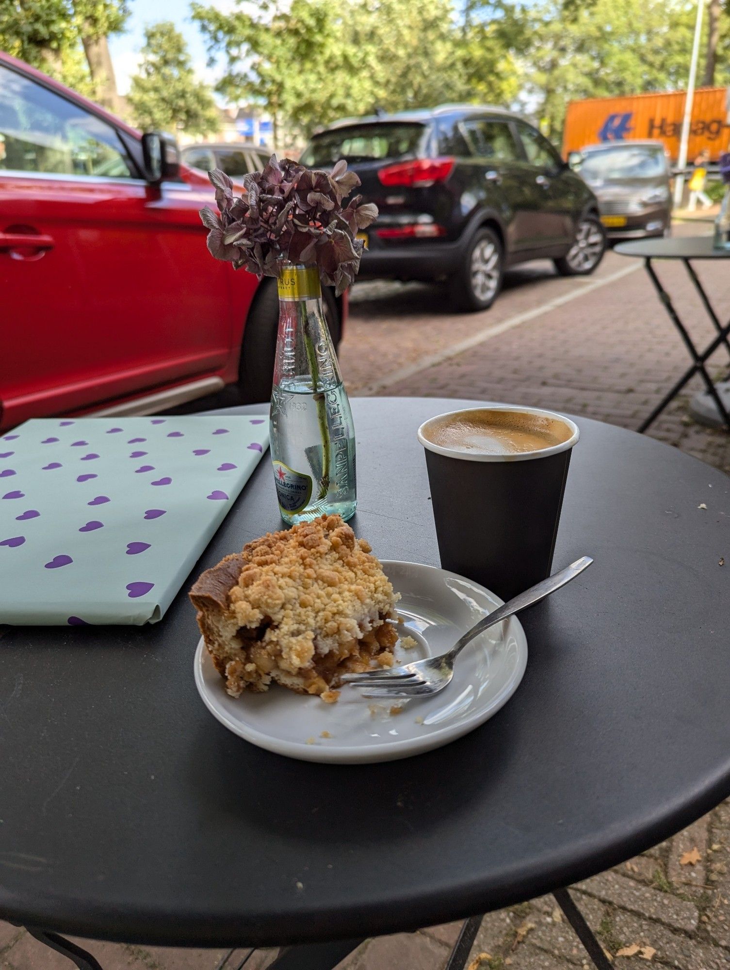 Een tafeltje met daarop een papieren bekertjes met koffie, een bordje met een enorme punt appel-crumble en een prentenboek ingepakt in groen papier met paarse hartjes. En een frisdrank flesje gevuld met water en paarse bloemen