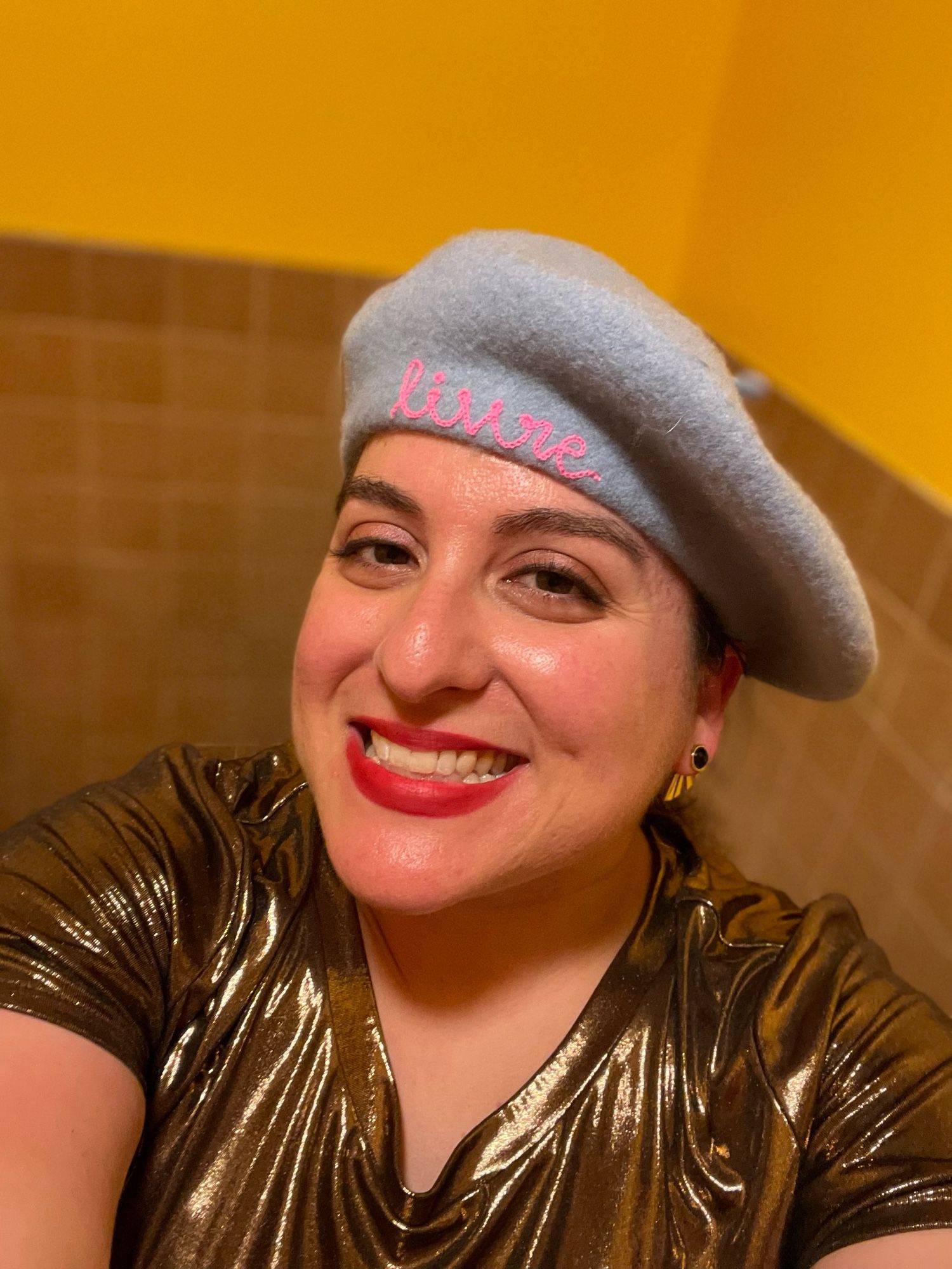 A picture of me, a white woman wearing a bronze metallic top and a periwinkle felt beret with the word “livre” embroidered on pink thread.