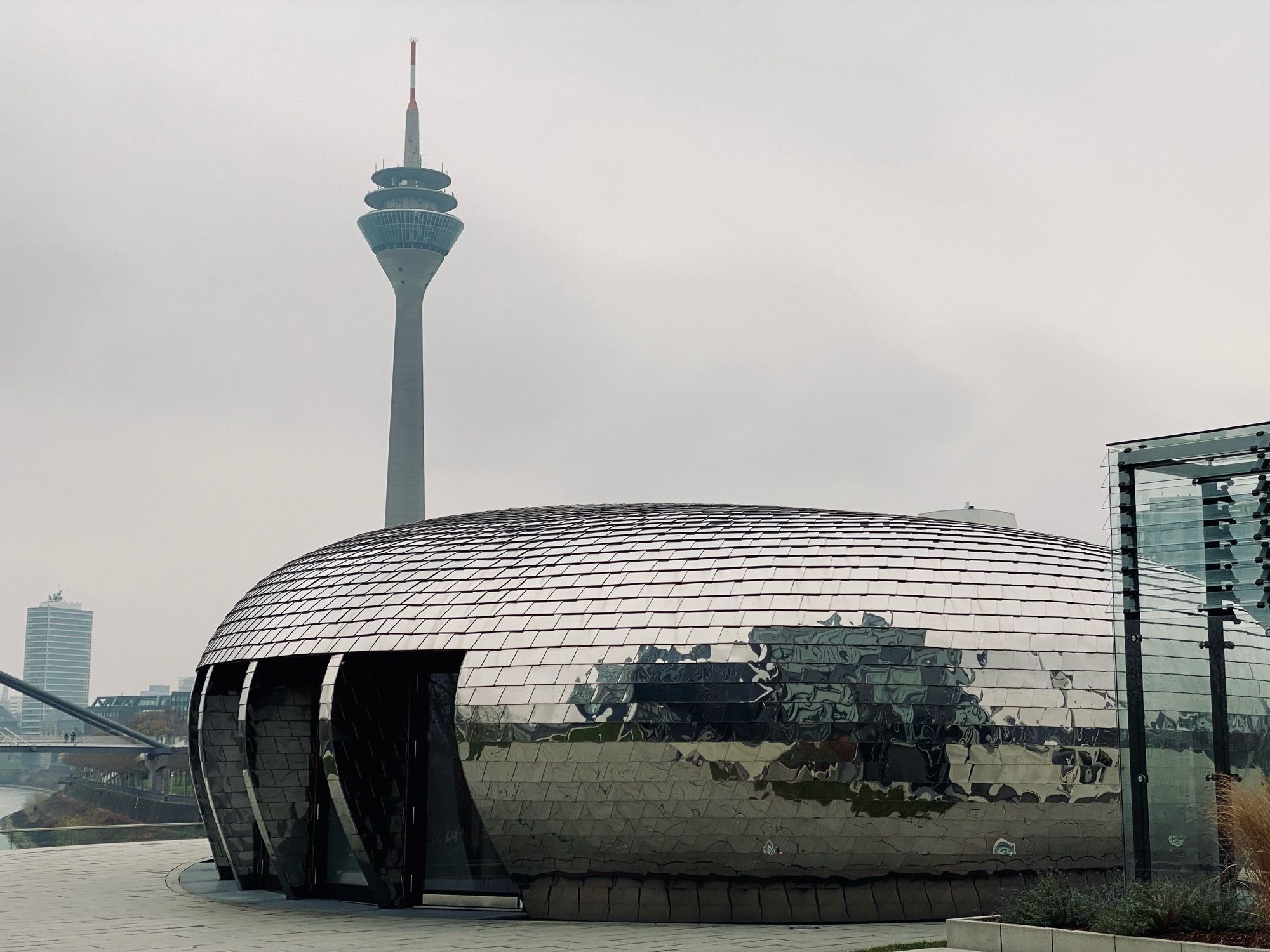 Im Hintergrund der Düsseldorfer Fernsehturm, der Rheinturm, am
linken Bildrand erkennt man ein Teil einer Brücke, dahinter ein Hochhaus. Im Vordergrund steht ovales Gebäude, das völlig verspiegelt ist. Darin spiegelt sich die Umgebung. Das Gebäude hat links mehrere bodentiefe, zurück versetzte Fenster. Ganz rechts sind Teile eines gläsernen Gebäudes sichtbar.