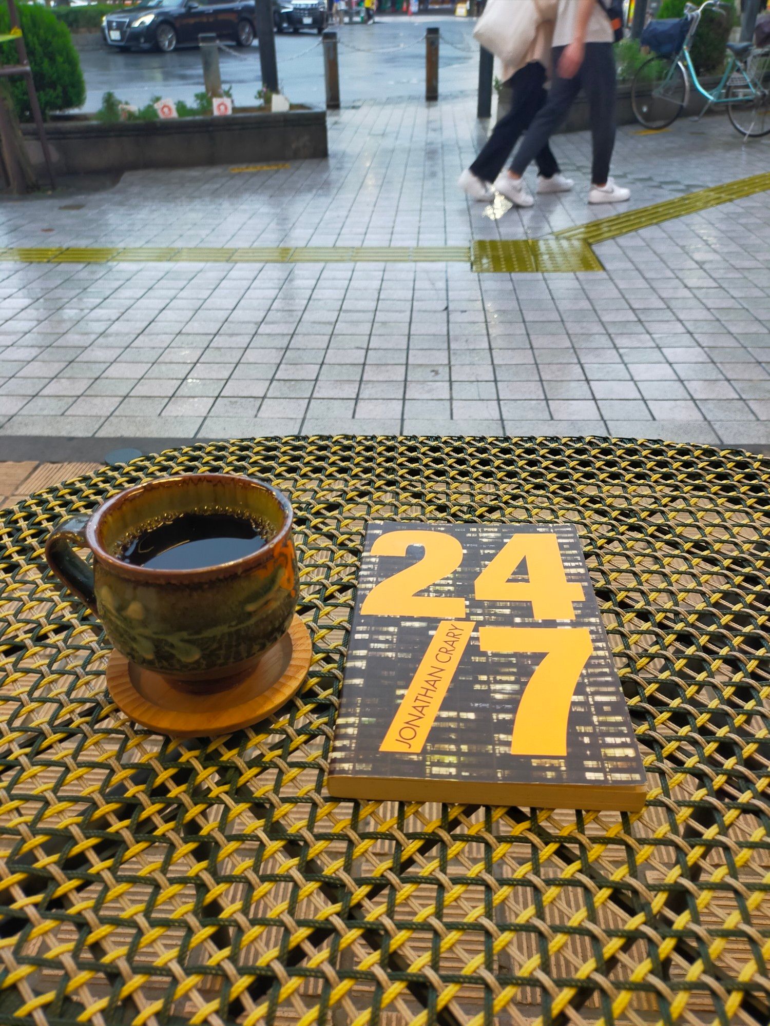 Photo is of a rainy day outside cafe seating area. The table is of woven gold fabric. The paperback book is on the table. There is a photo of a nighttime apartment building and very large 24/7 in bold gold font overlaying the photo. The gold of the book letters somewhat matches the gold woven table. To the left is a brown mug of black coffee on a sandstone round cosster. In the distance you can see the wet gray tiles of the walkway and the yellow rubber tactile tiles for the blind intersecting across. You can see the jeaned legs and white sneakered feet of 2 people walking by