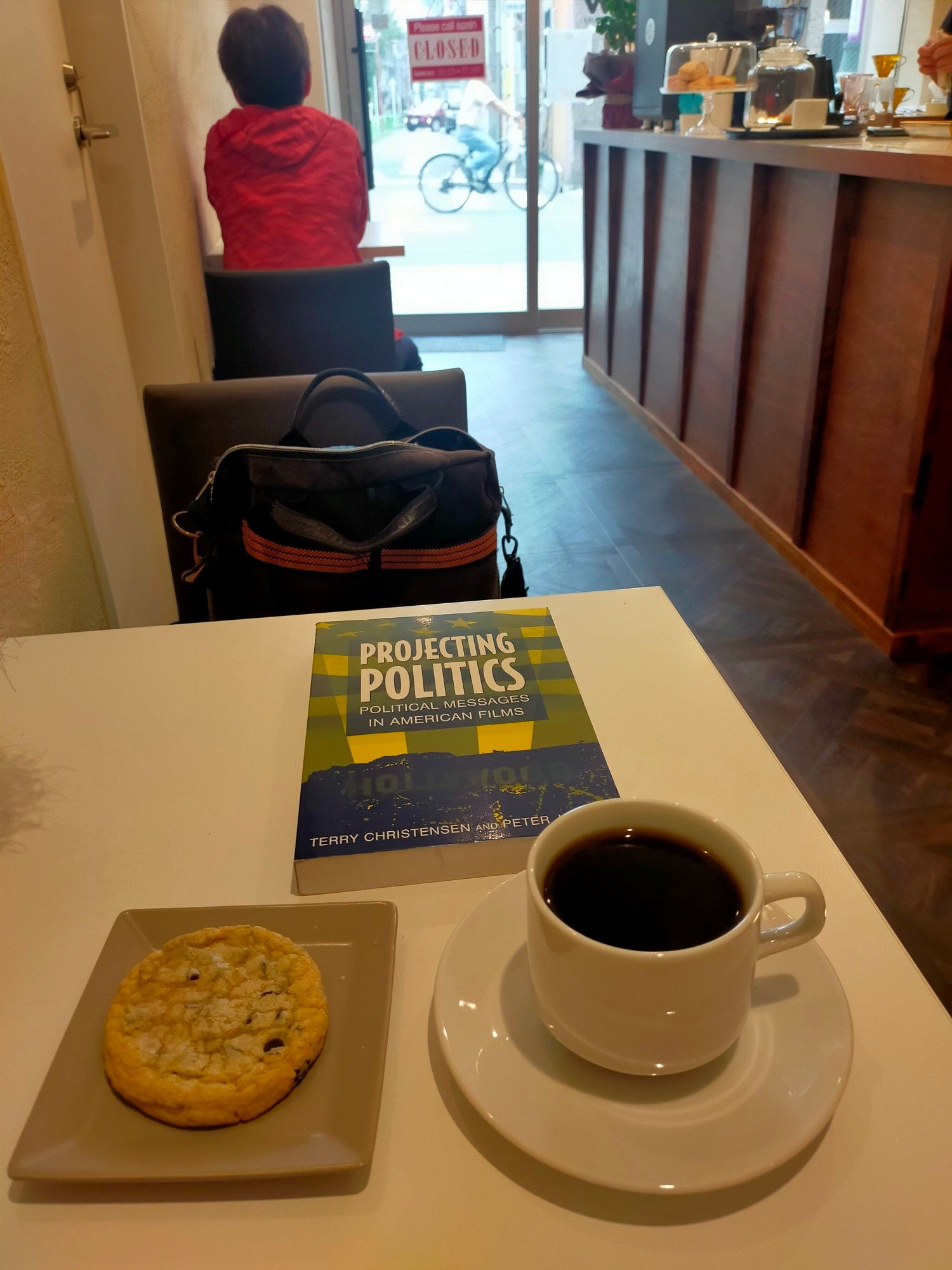 Photo is of the inside of the cafe. In the foreground is a white table with a white mug & saucer of black coffee. To the left is a small square grey plate with a chocolate chip cookie on it. Centered above both is the paperback book PROJECTING POLITICS. In the chair opposite is a brown shoulder bag. Further back is the back of a customer sitting. She is wearing a red hoodie & has short hair. In the furthest distance outside the cafe glass door, a blurry cyclist heading right can be seen on the street. To the right of the tables, the brown counter can be seen.