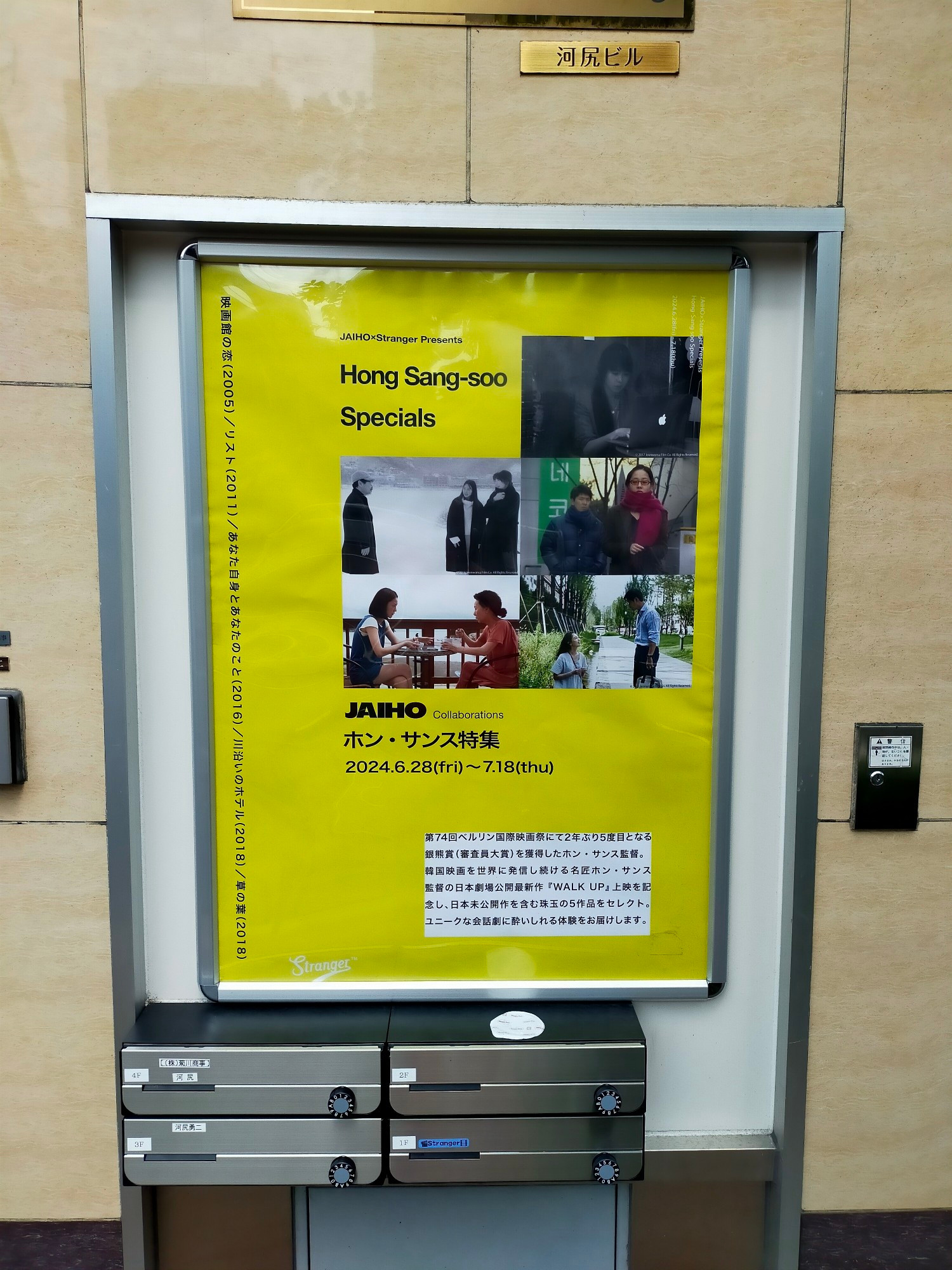 Photo of a yellow movie poster outside the venue. It reads (misspelling the director's given name) "Hong Sang-soo Specials". On the poster are stills from 5 HONG Sangsoo films Bottom right is from YOURSELF AND YOURS where a Korean man is looking down on a Korean woman in a grassy stream. Above that is from TALE OF CINEMA with a Korean woman in a purple scarf & sunglasses being followed by a Korean man on a sidewalk. Above that is a black and white still of a Korean woman in an unindentifiable Hong film. I also can't identify the films on the left, top features a bespectacled Korean man looking at 2 Korean woman who look back and below a young Korean woman sits at an outside table across from an older Korean woman