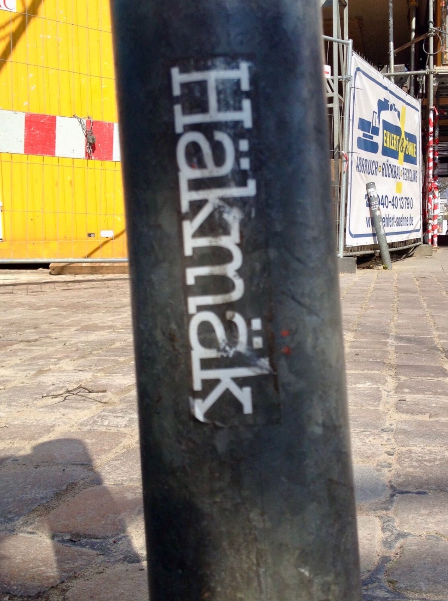 Schiefer Pfahl in einer Stadt, auf dem ein Sticker klebt, wo „Häkmäk“ drauf gedruckt ist.