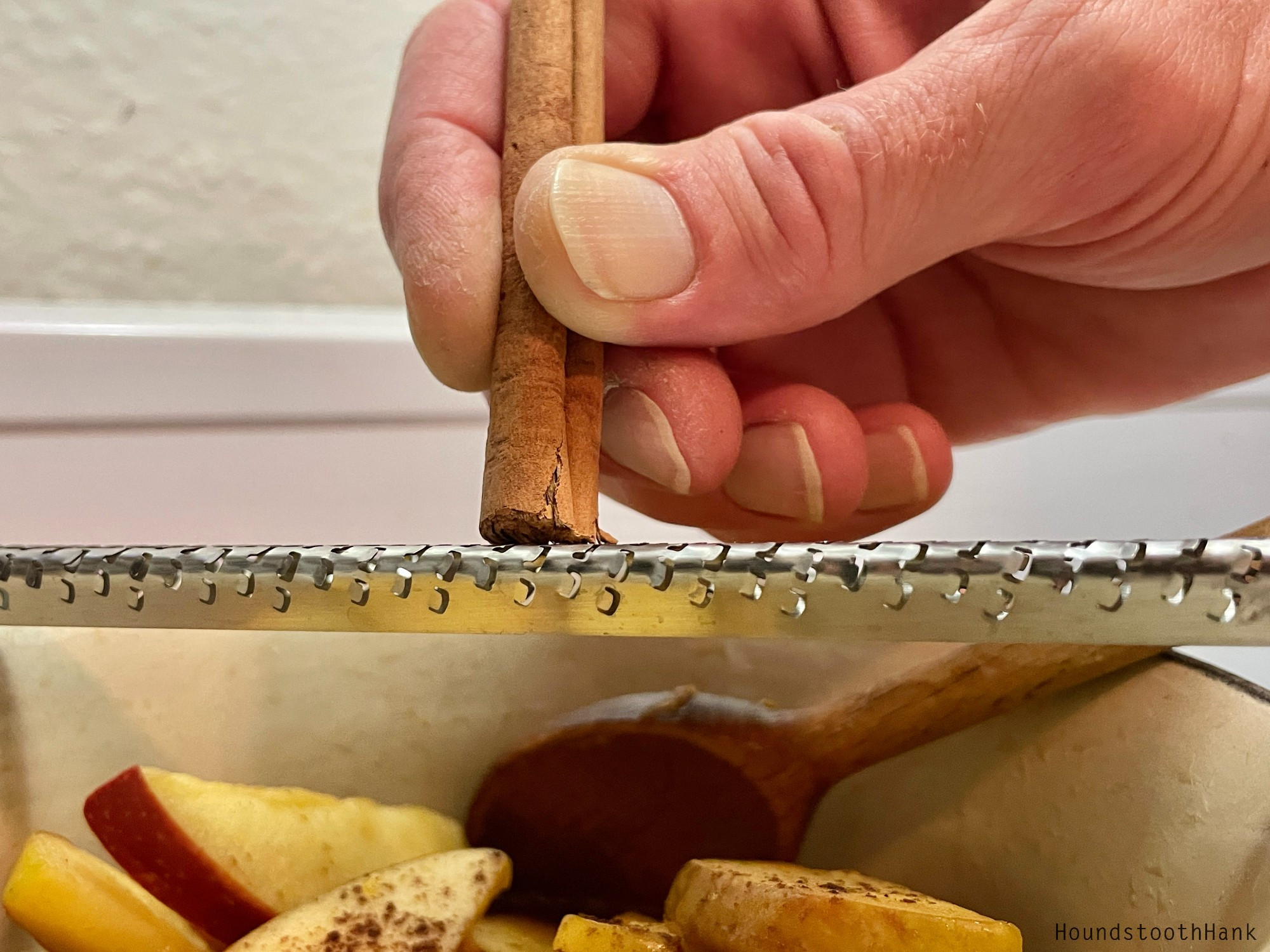 Closeup of Hank's fingers dragging a stick of cinnamon across a microplane over a bowl of sliced apples