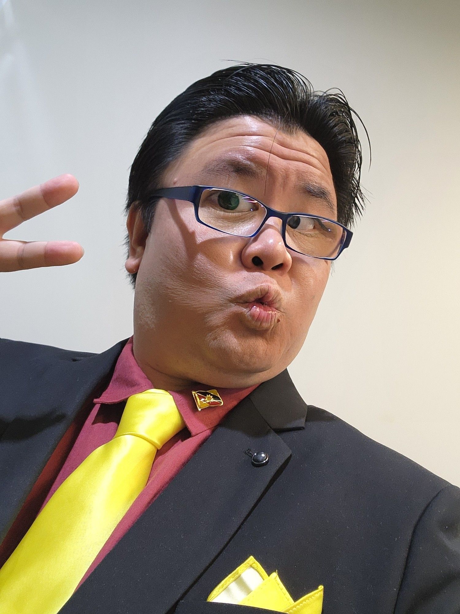 Photo selfie of myself in a black suit, red shirt, and yellow tie - mimicking the colors of the Sarawak flag, whose pin that belonged to my late dad is attached to my lapel. I am doing the duck face while throwing up V fingers