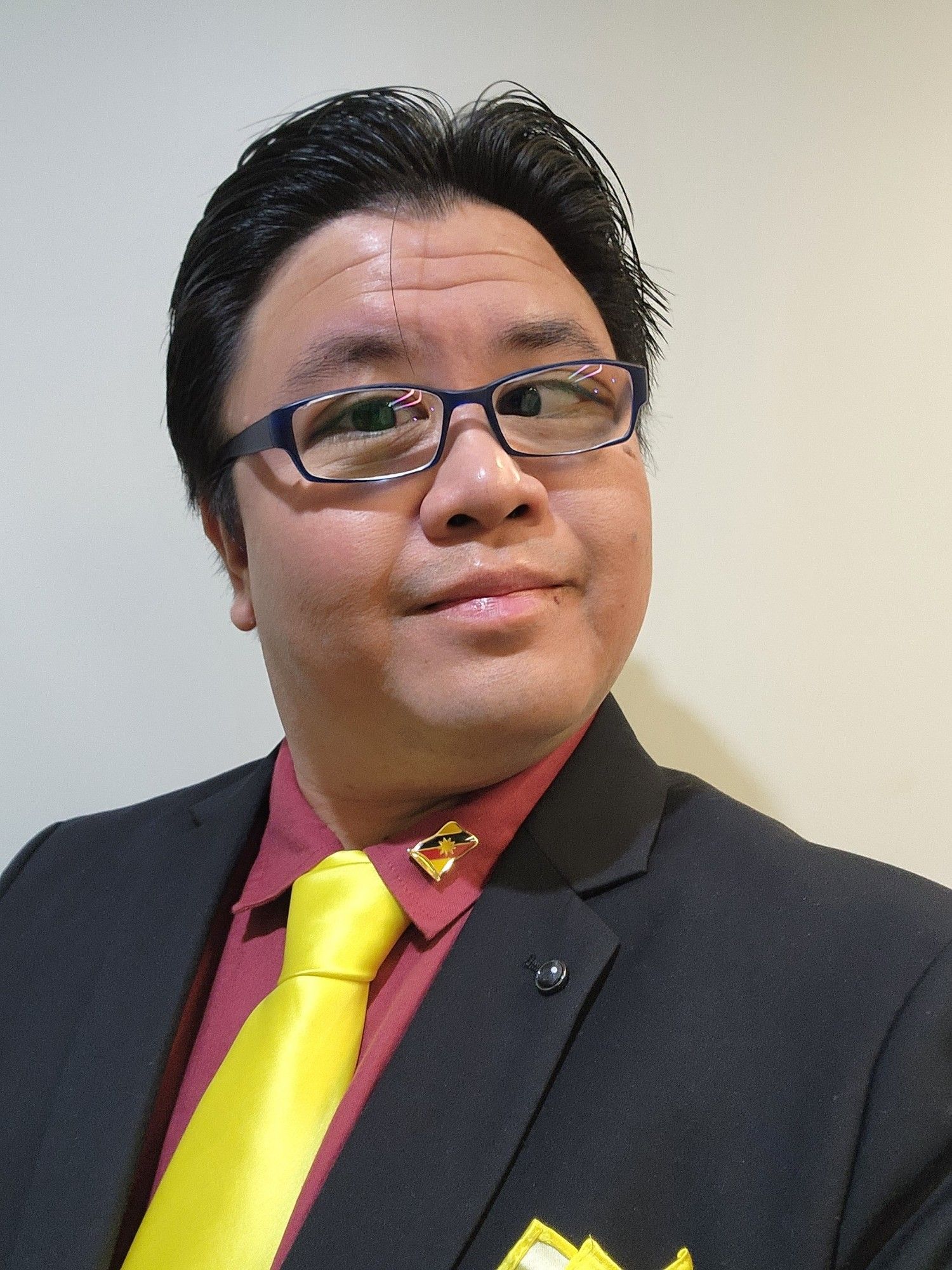 Photo selfie of myself in a black suit, red shirt, and yellow tie - mimicking the colors of the Sarawak flag, whose pin that belonged to my late dad is attached to my lapel. I have a fairly friendly expression