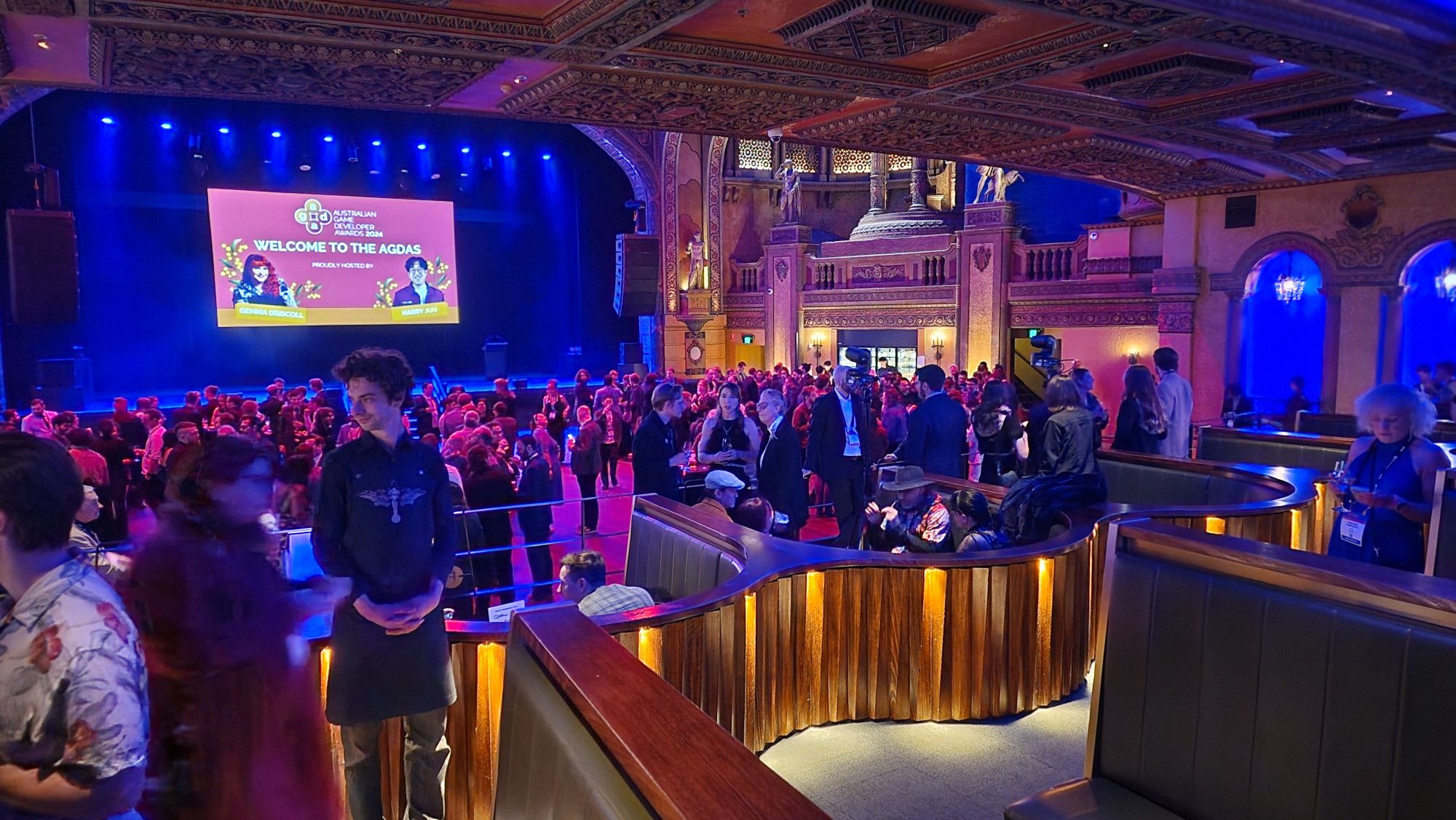 A photo of the Forum from further back, lit up and done up to the nines for the AGDAs 2024. There is a massive crowd of people milling in the dance floor area in the background in front of the big screen and podium where the presentation will take place and awards handed out. Foreground has booths where the special guests/sponsors of that night's show would be seated