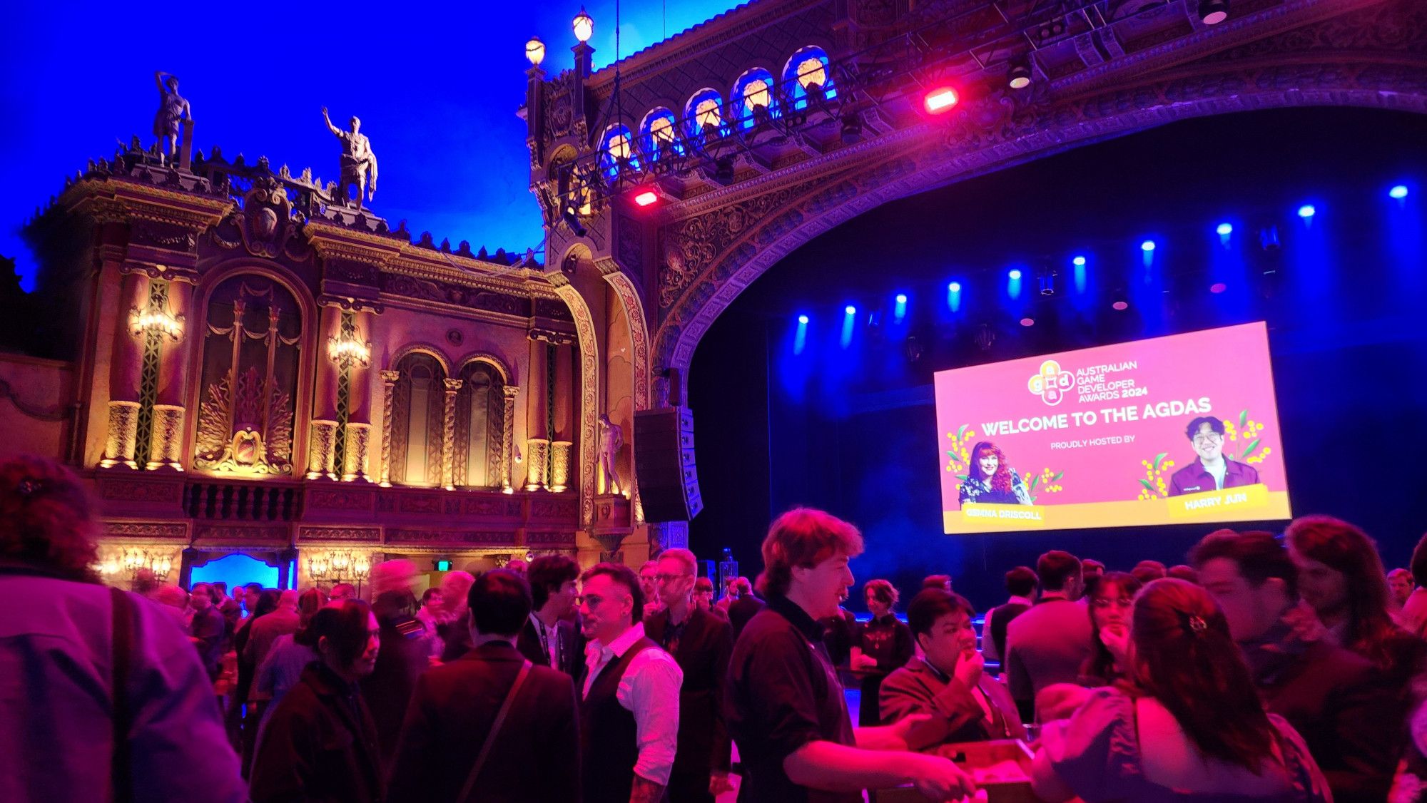 A photo of the Forum from the open dance floor area, lit up and done up to the nines for the AGDAs 2024. There is a massive crowd of people milling in front of the big screen and podium where the presentation will take place and awards handed out. Fancy mouldings done up with Greek/Roman(esque) statues dominate the outer edges of the stage