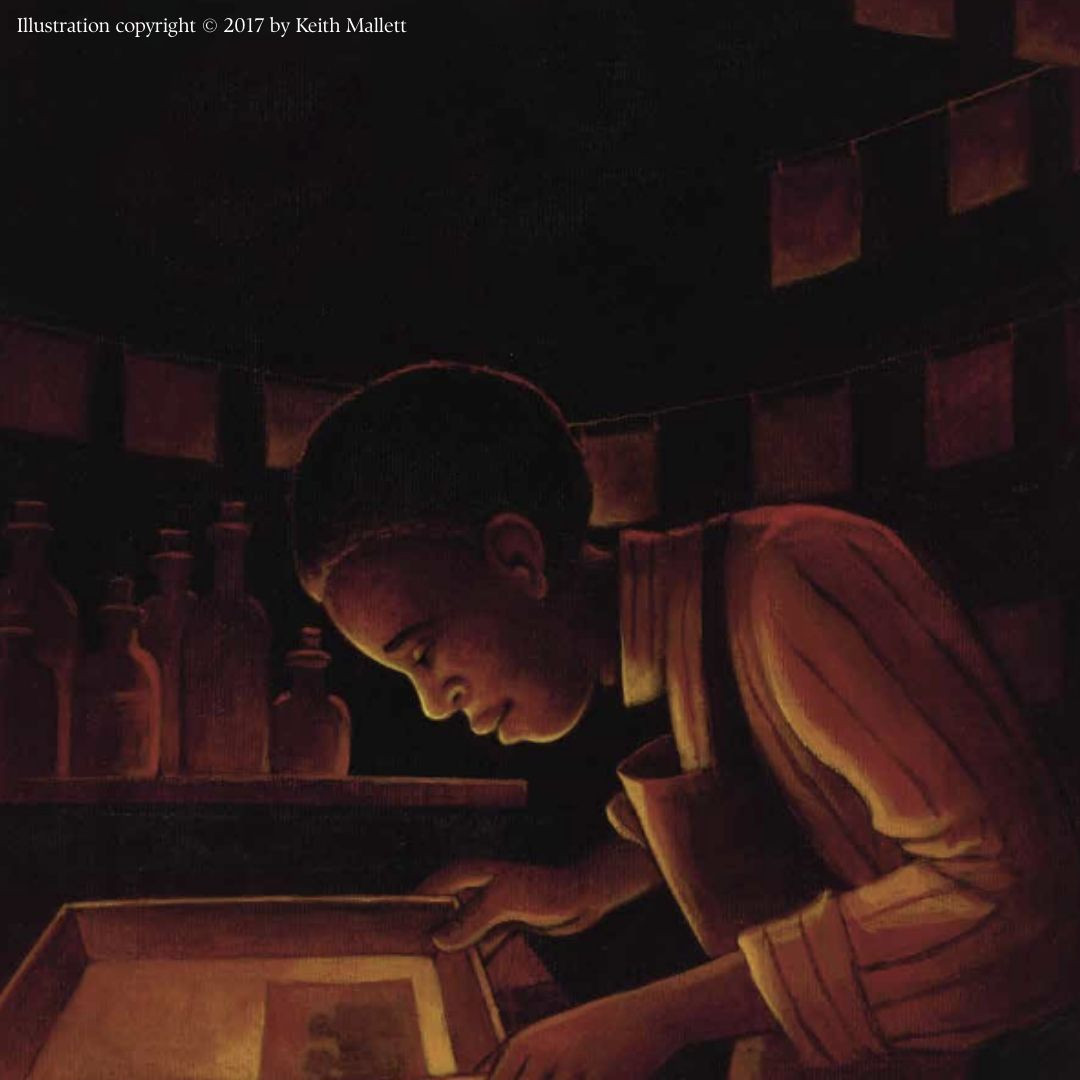 Excerpt page from the picture book biography, “Take a Picture of Me, James VanDerZee!” by Andrea J. Loney, illustrated by Keith Mallett. The illustration depicts young James VanDerZee in a dark room developing a picture. Illustration copyright © 2017 by Keith Mallett.