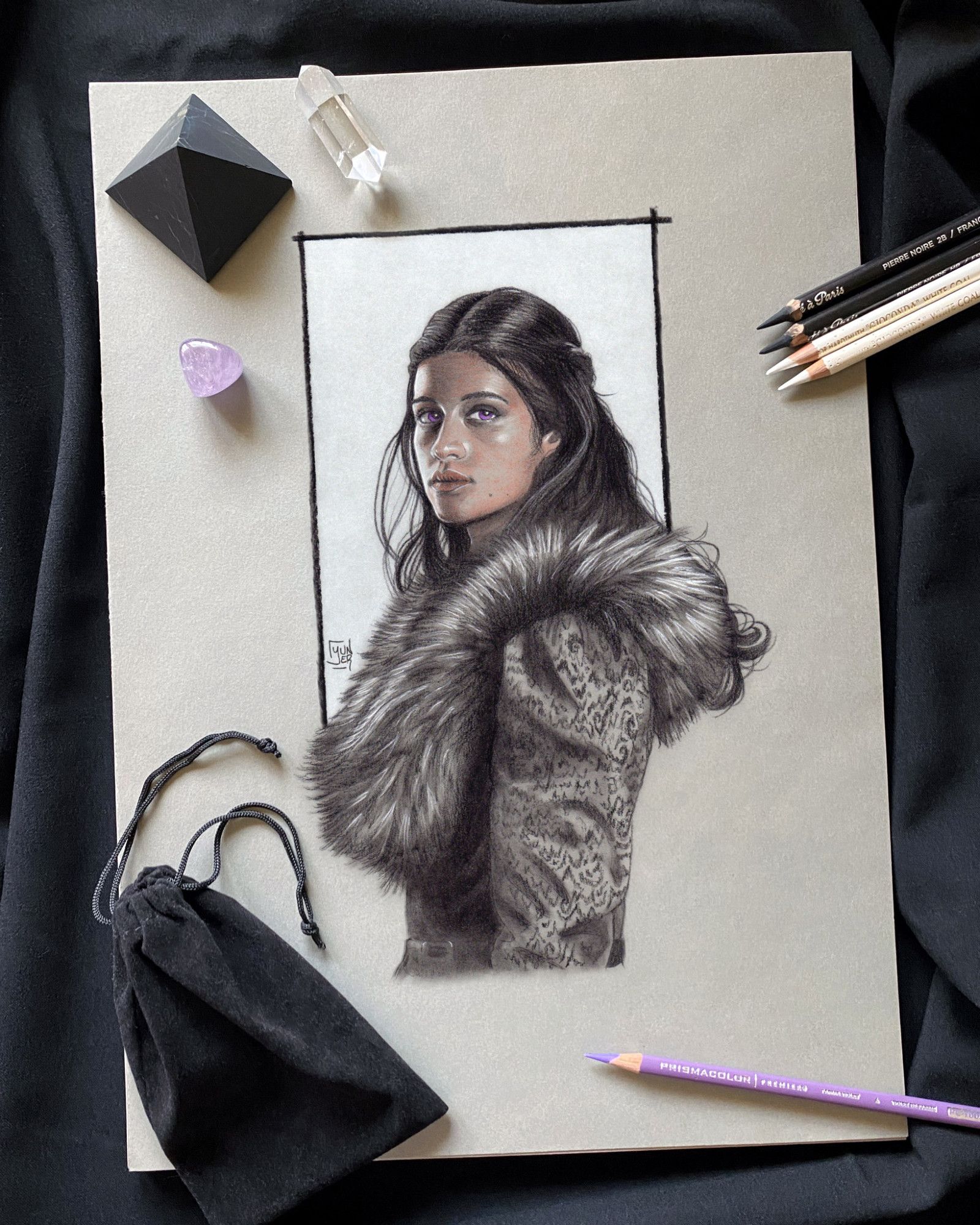 Portrait of Yennefer of Vengerberg (Anya Chalotra) from a scene of "The Witcher", made with pierre noire, sanguine and white chalk on grey toned paper; around the drawing on the table there are pencils (used for the drawing), some crystals with a black stone pyramid, and a small black velvet bag.