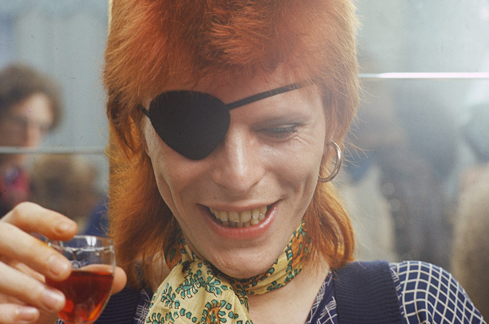 David Bowie in an eyepatch and scarf drinking some sort of dessert wine.