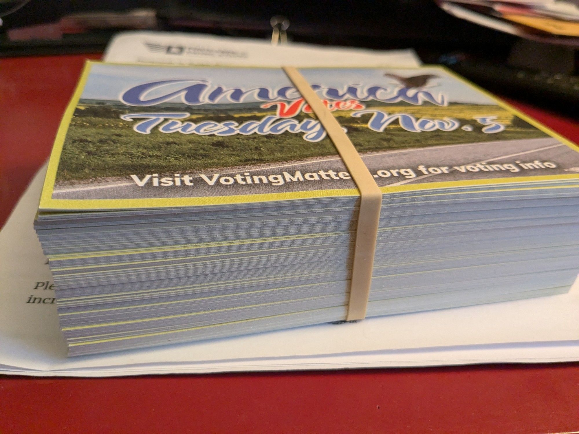 A stack of 150 postcards to swing states held together by a rubber band. Image on the postcard says America Votes Tuesday November 5, visit VotingMatters.org for voting info.