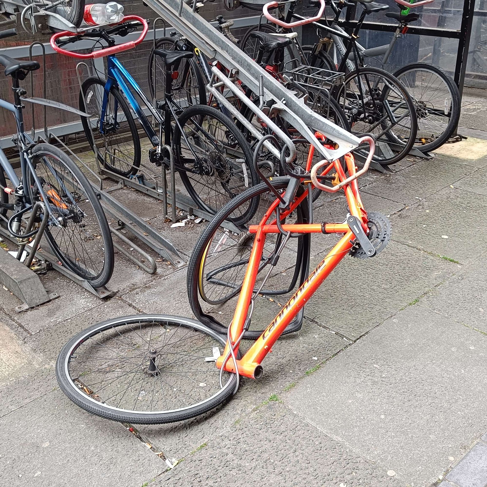 Red bike frame attached to extended tray of two tier bike parking, in poor condition with wheels hanging on by lock & cables & no handlebars