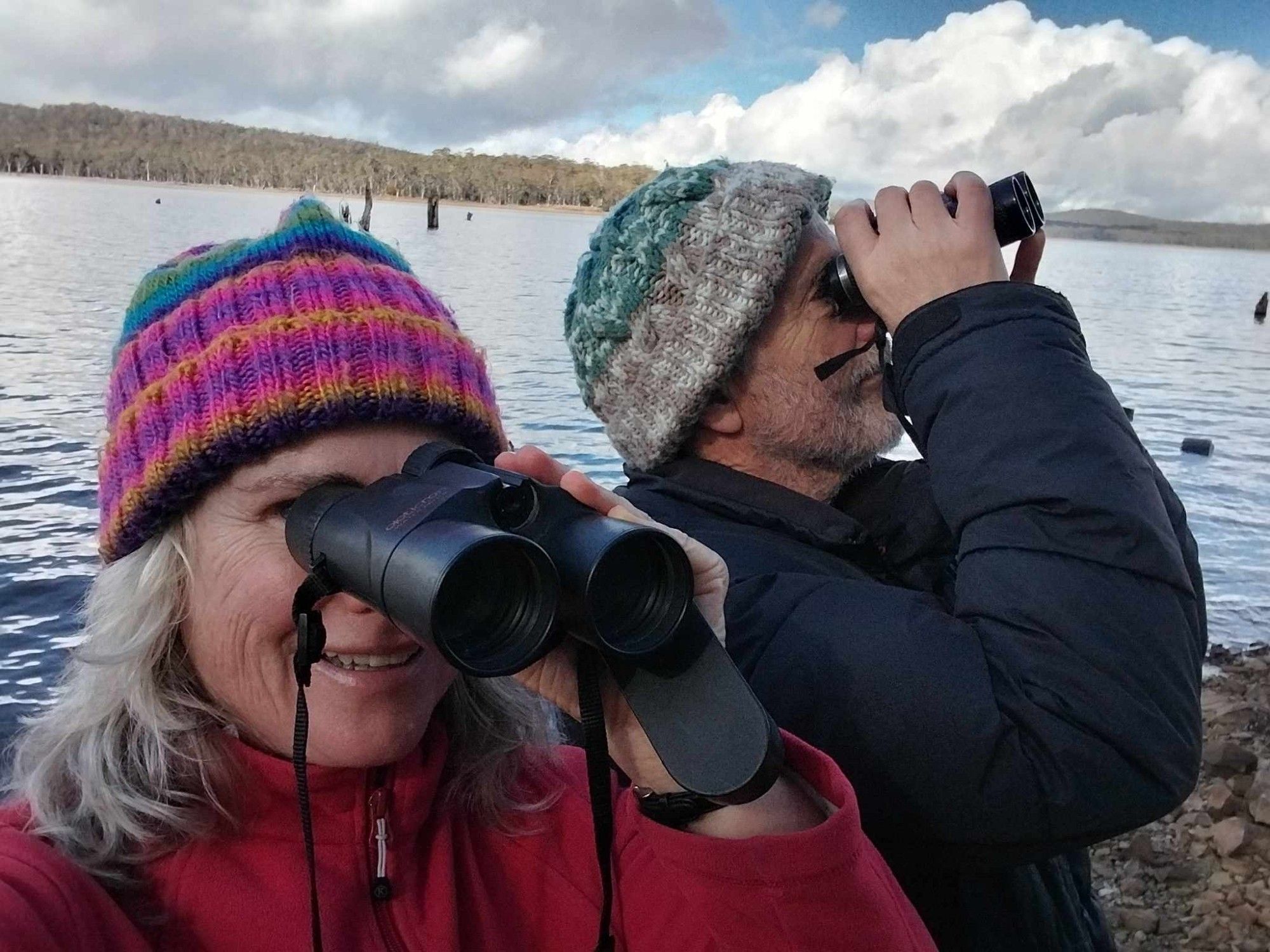 Two people wearing warm outdoor clothing, including colourful knitted beanies, with a lake behind them, look in two different directions with their binoculars. Photographed by Chris Grove.