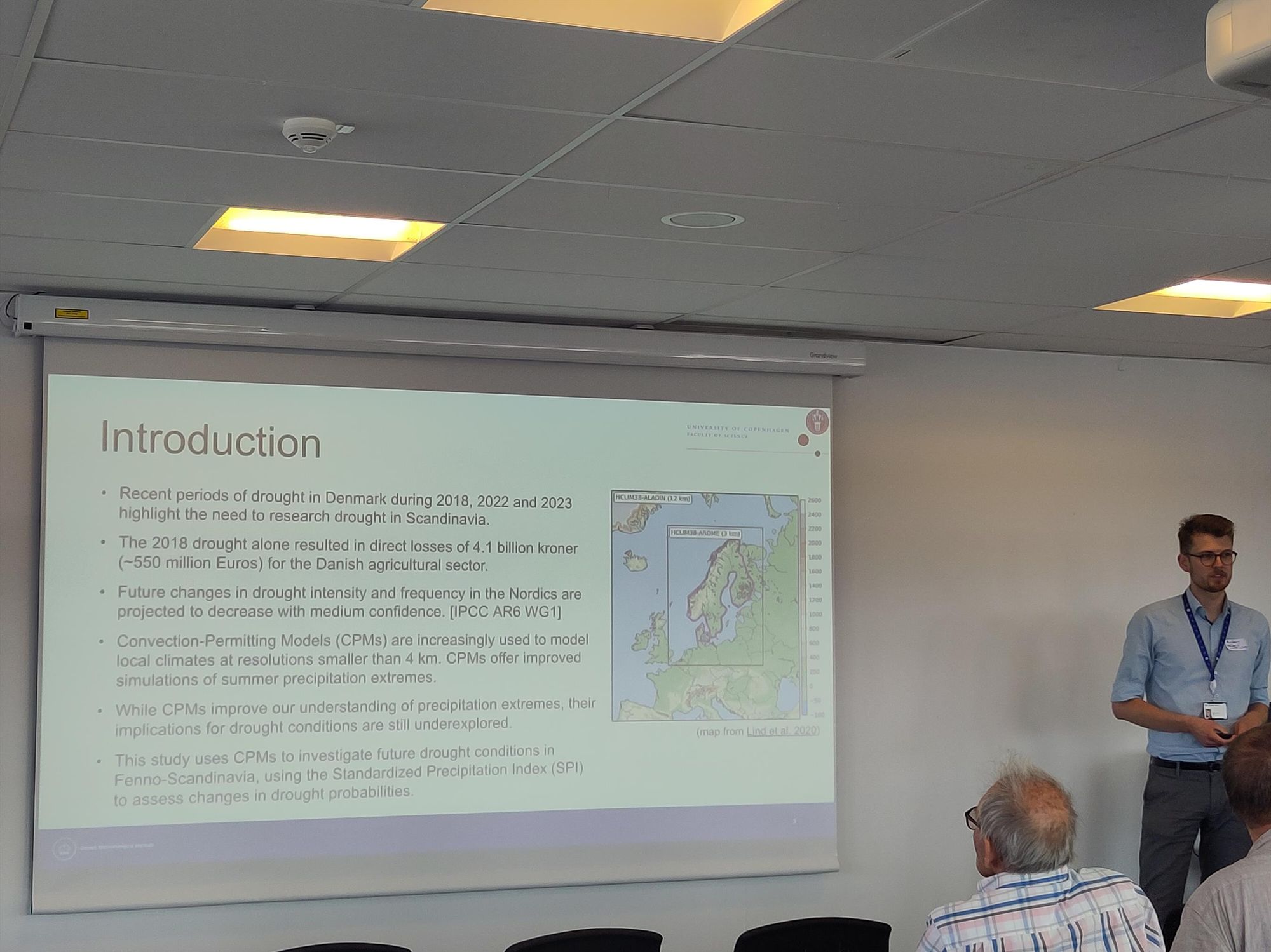 A young man stands in front of a slide summarising a study outline 