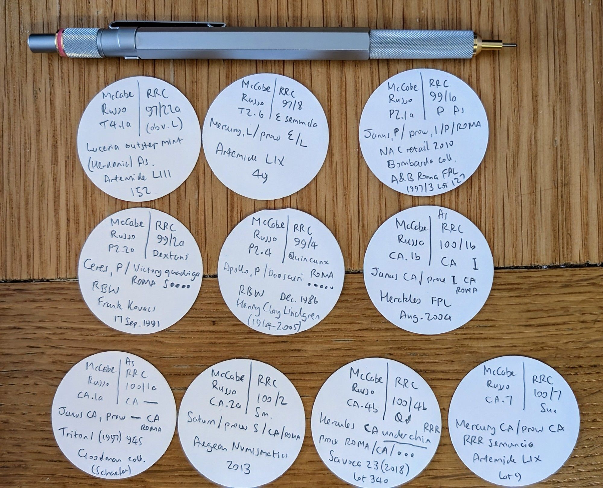 Rotring 800 metal mechanical pencil and 10 hand written round tickets describing ancient coins each about 40mm diameter, on a wooden table surface.