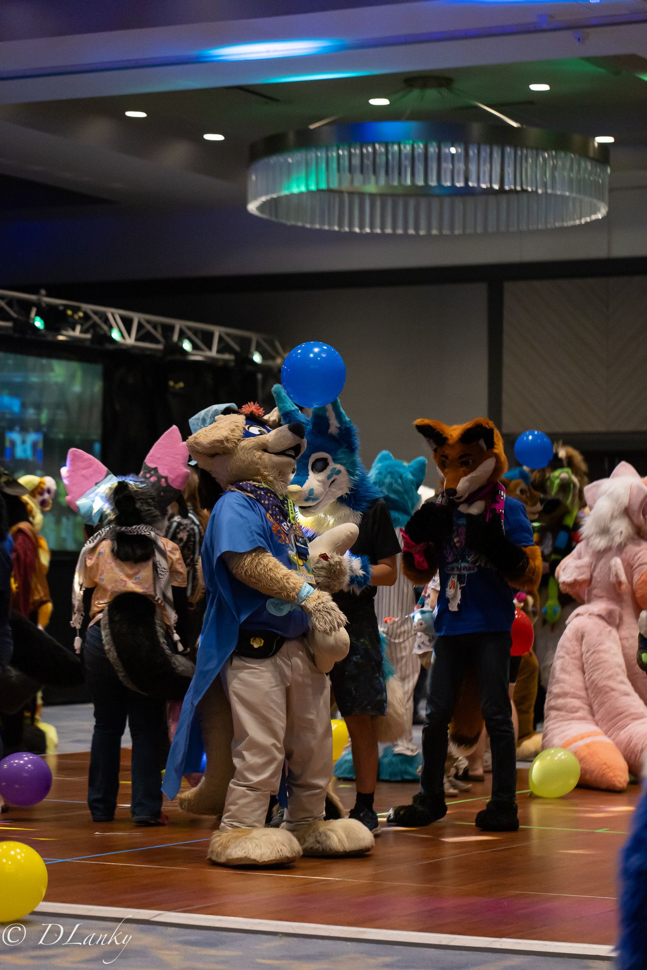 A bunch of fursuiters stand on a dance floor, surrounded by balloons.  RK, a yellow flop-eared superhero dog, tries no nose a blue balloon floating above him.