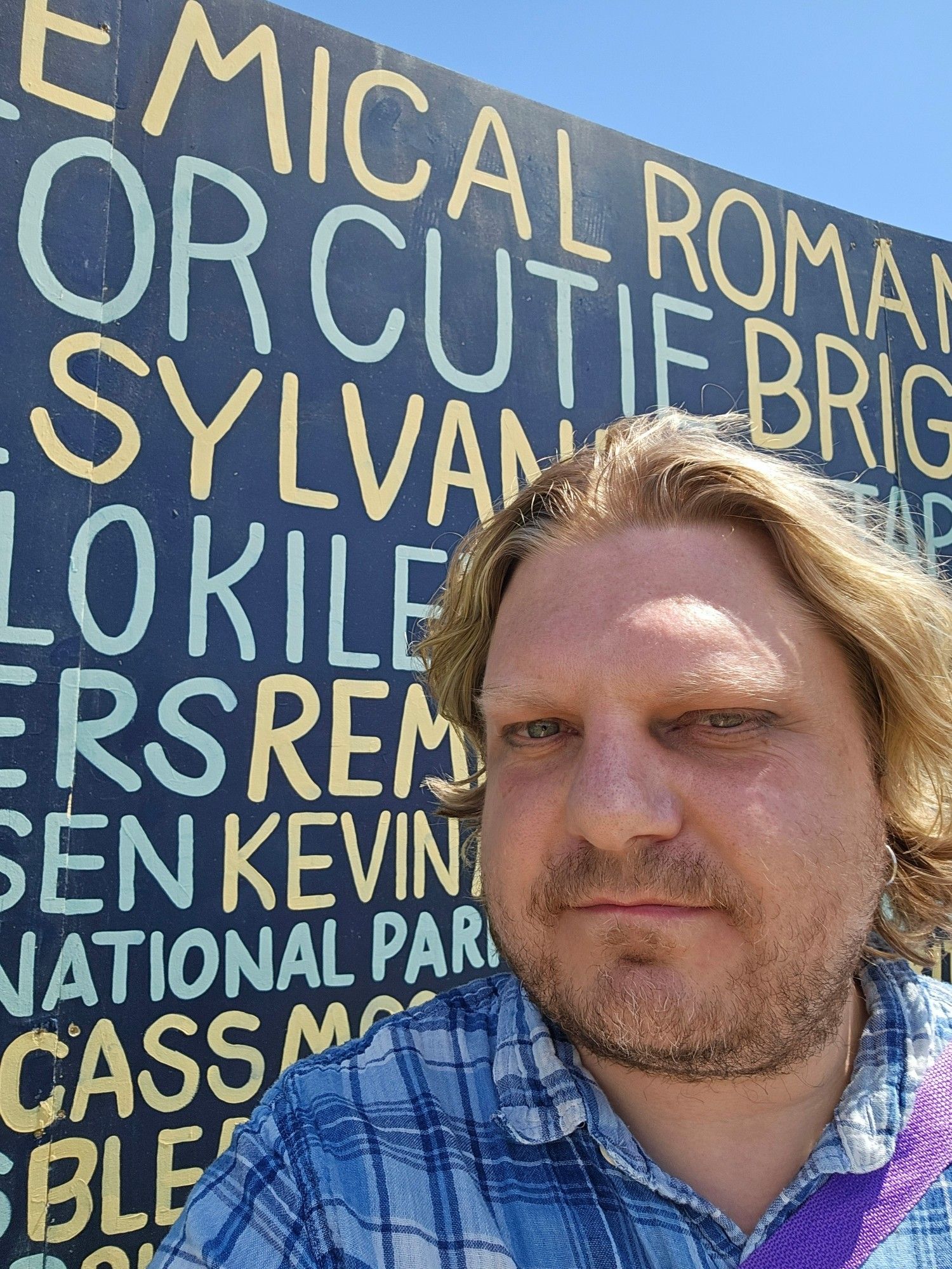 Jeremy in a plaid shirt, long hair down in front of the Kilby Wall of Dame listing artust names. Rilo Kiley is prominently featured.