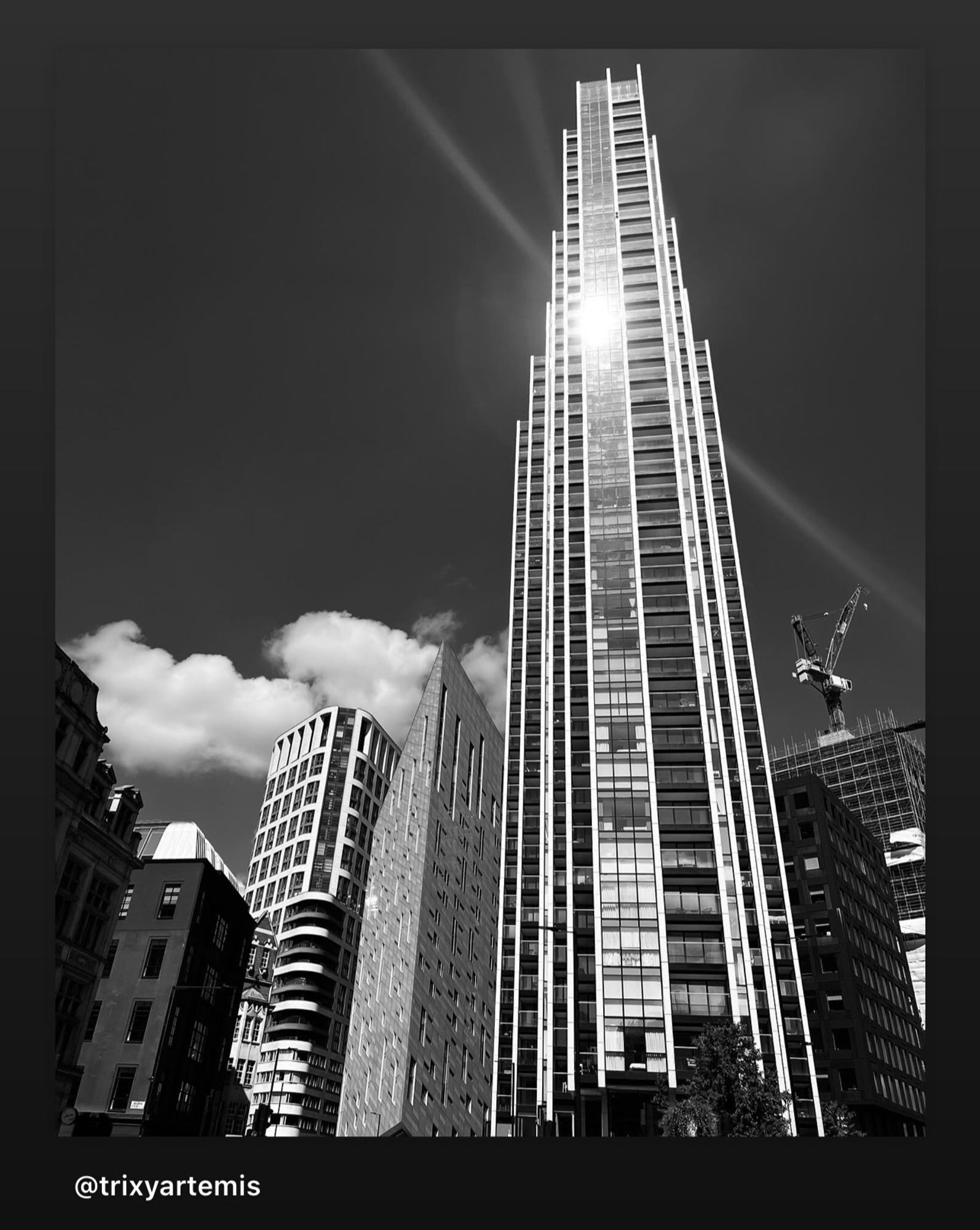 Black and white photograph of a skyscraper full glass windows all the way up and the building gets narrower as it gets higher and the sun is reflected in its windows creating sharp sun rays that radiate outwards! The Montcalm east is next to the building and creates an optical illusion in its structure.