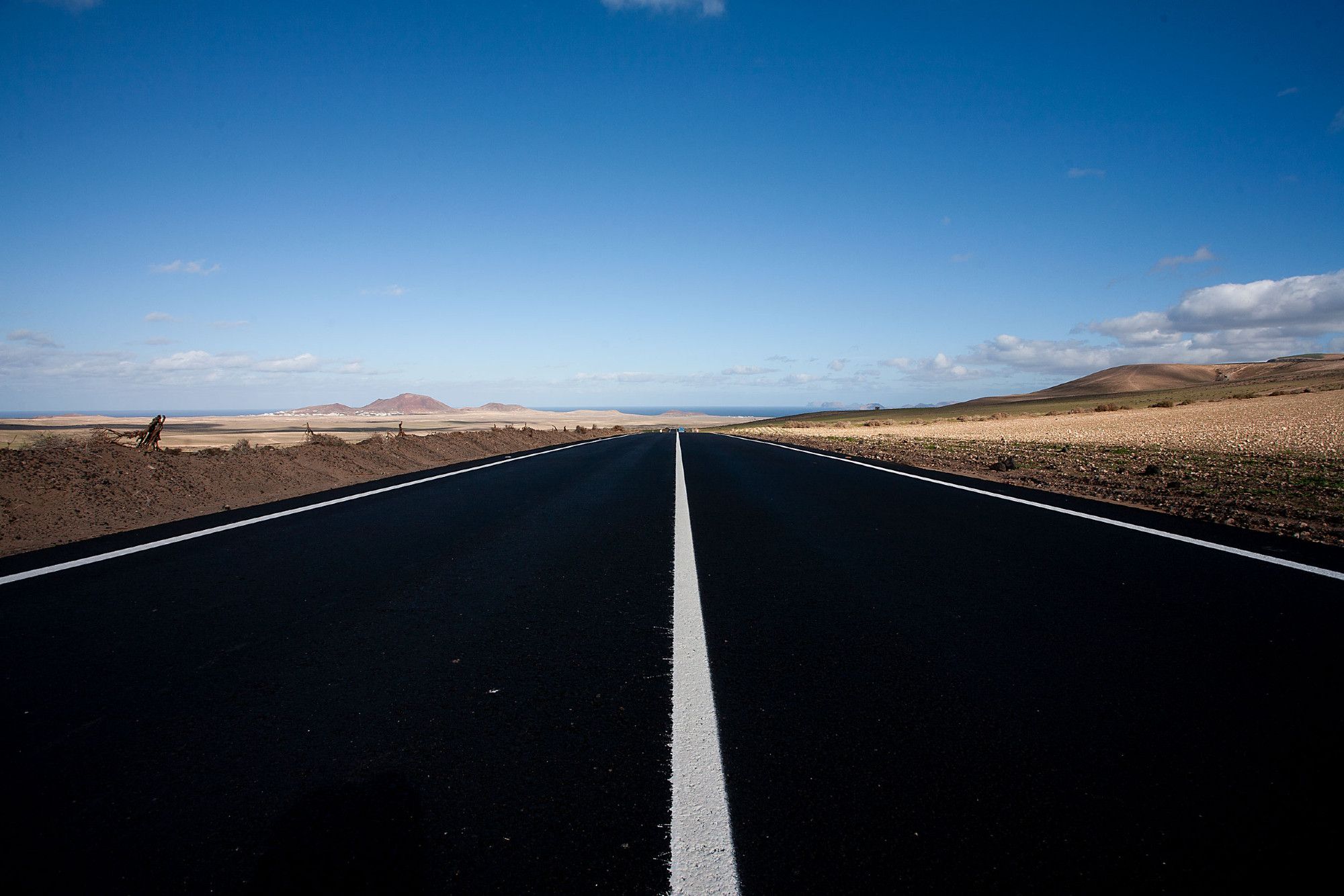 Eine Straße auf Lanzarote, die scheinbar in die Unendlichkeit führt. Blauer Himmel, karge Landschaft rechts und links.