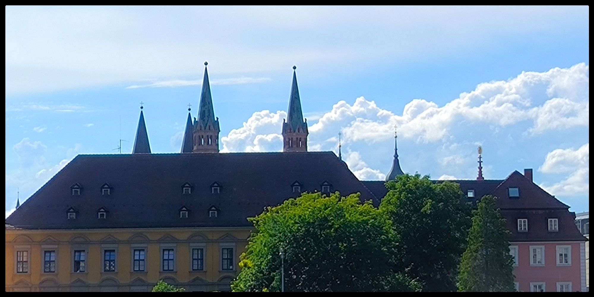 Blick auf ein Gebäude, vor dem ein Baum steht. Hinter dem Gebäude sind mehrere Kirchtürme zu sehen. Wolken bedecken teilweise den Himmel.