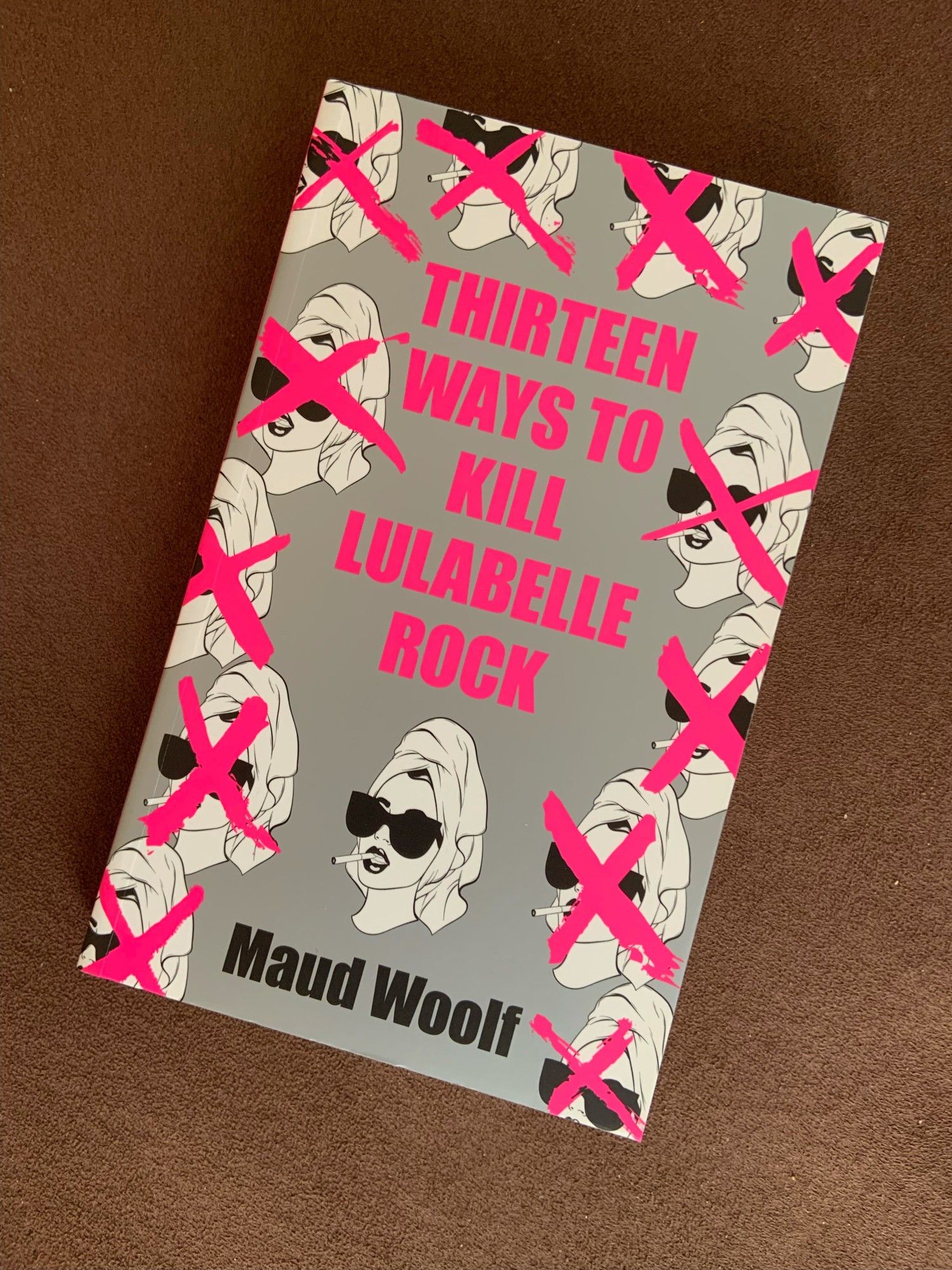 The cover of the novel "Thirteen Ways to Kill Lulabelle Rock" by Maud Woolf. It is chrome gray and hot pink, and features indentical images of the head of the title character, twelve of which are "x"ed out.