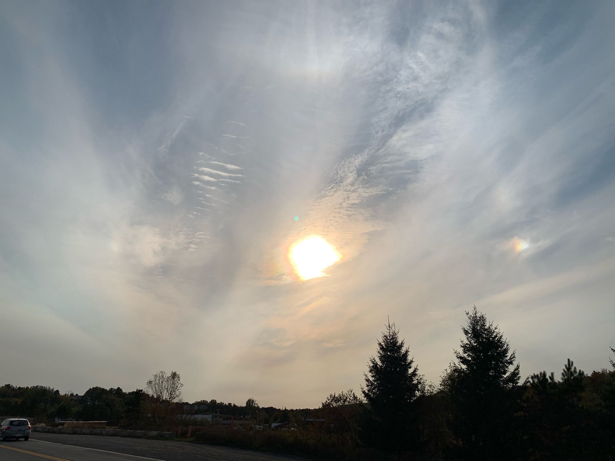 A faintly cloudy blue sky over a dark horizon. The sun is in the centre and a sun dog — a bright refracted image with a rainbow hue — is visible to the right. There’s even a suggestion of a halo around the sun.