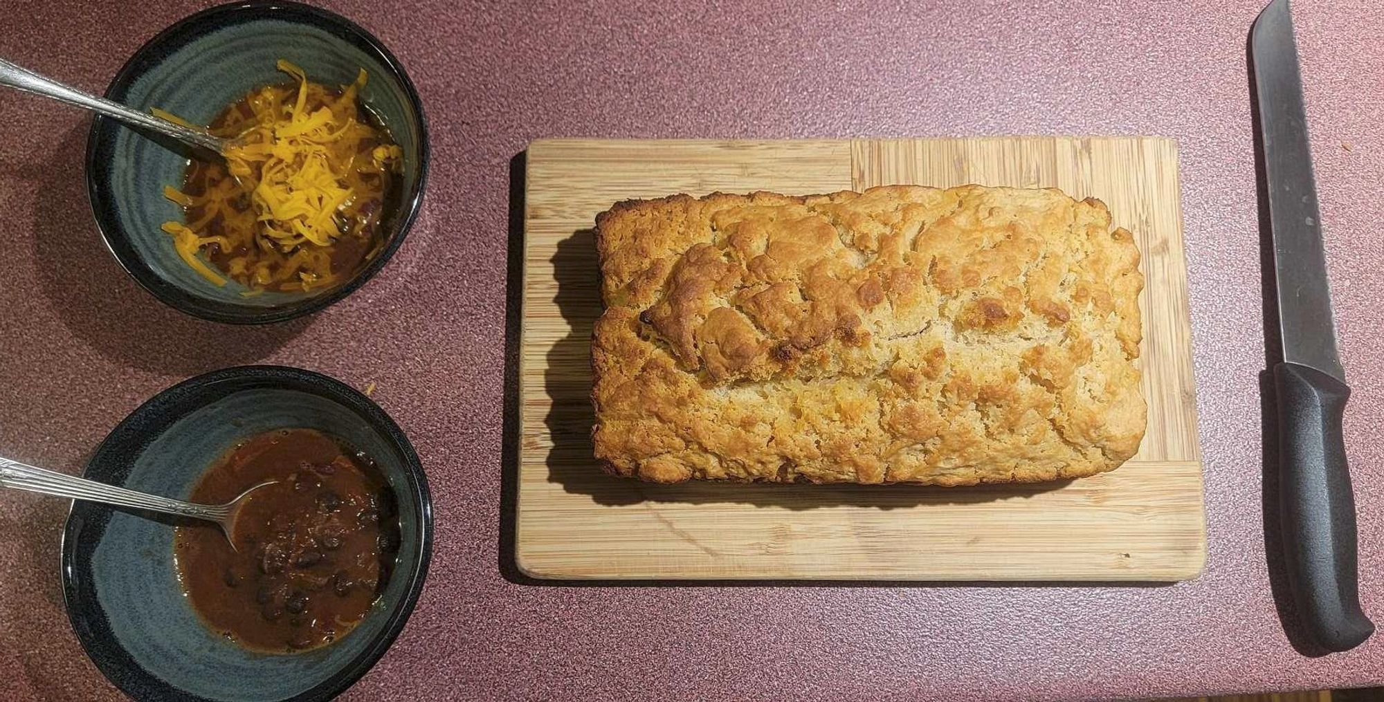 bread on a cutting board sided by two bowls of chili and a breadknife.