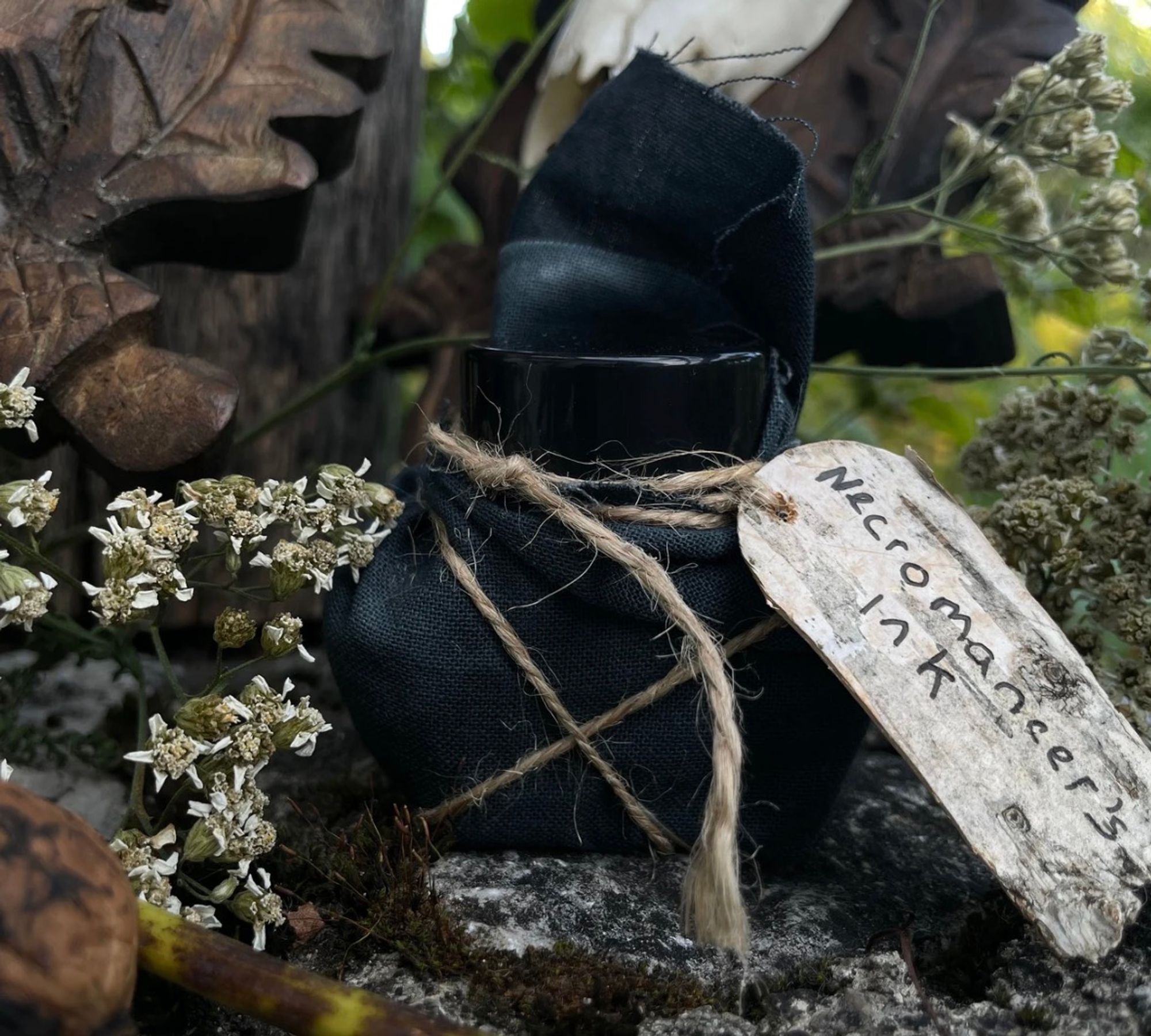 A bottle of Necromancer's Ink wrapped in black cloth sitting on stones and surrounded by flowers and moss.