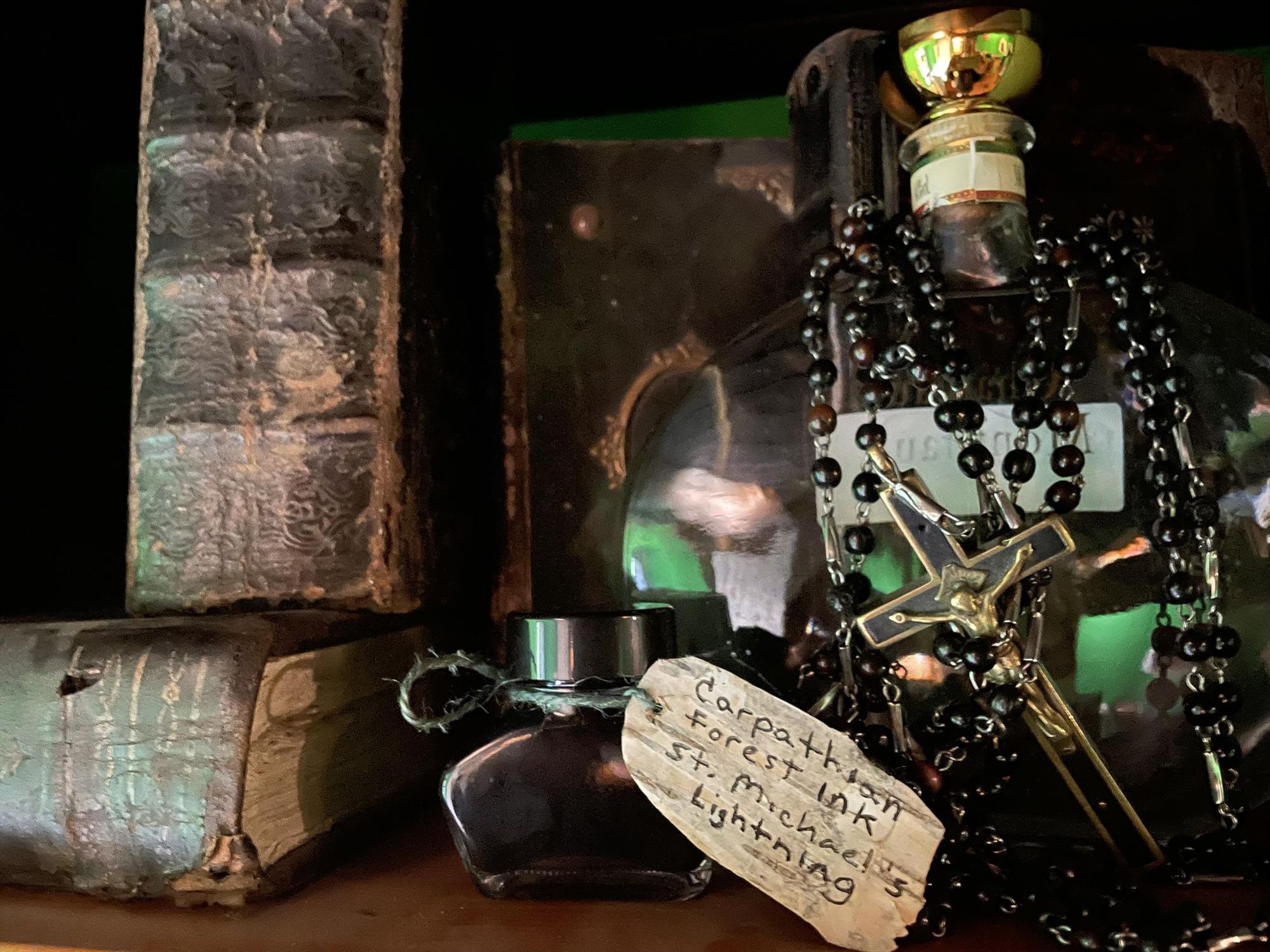 A small squat bottle of ink labelled "Carpathian Forest Ink St. Michael's Lightning" sitting on a shelf amidst books, and a large empty bottle draped with a necklace featuring a cross.