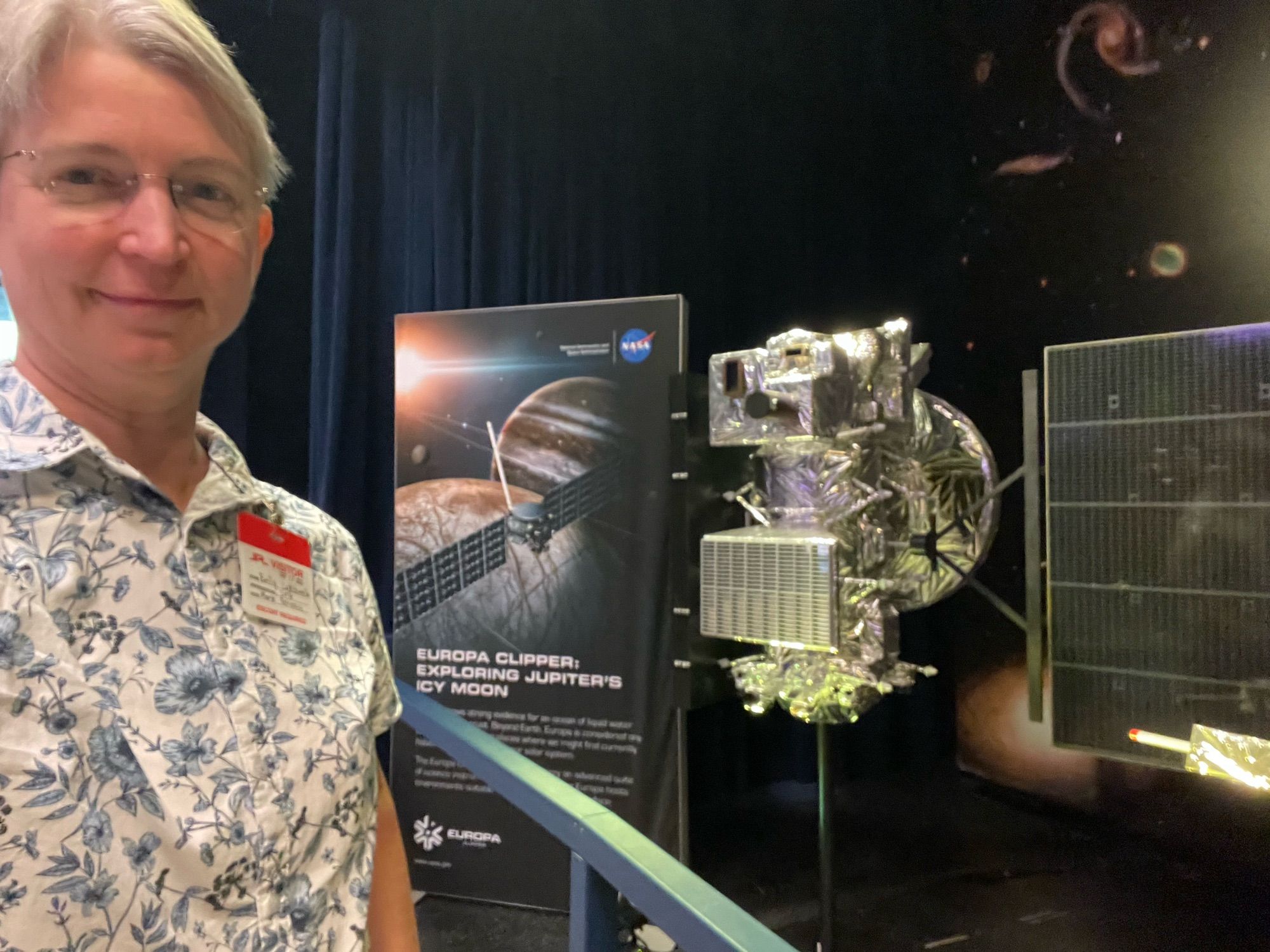 Selfie taken in front of a 1/4 scale model of the Europa Clipper spacecraft, a lumpy box wrapped in silver foil with solar panels that are large even at this scale.