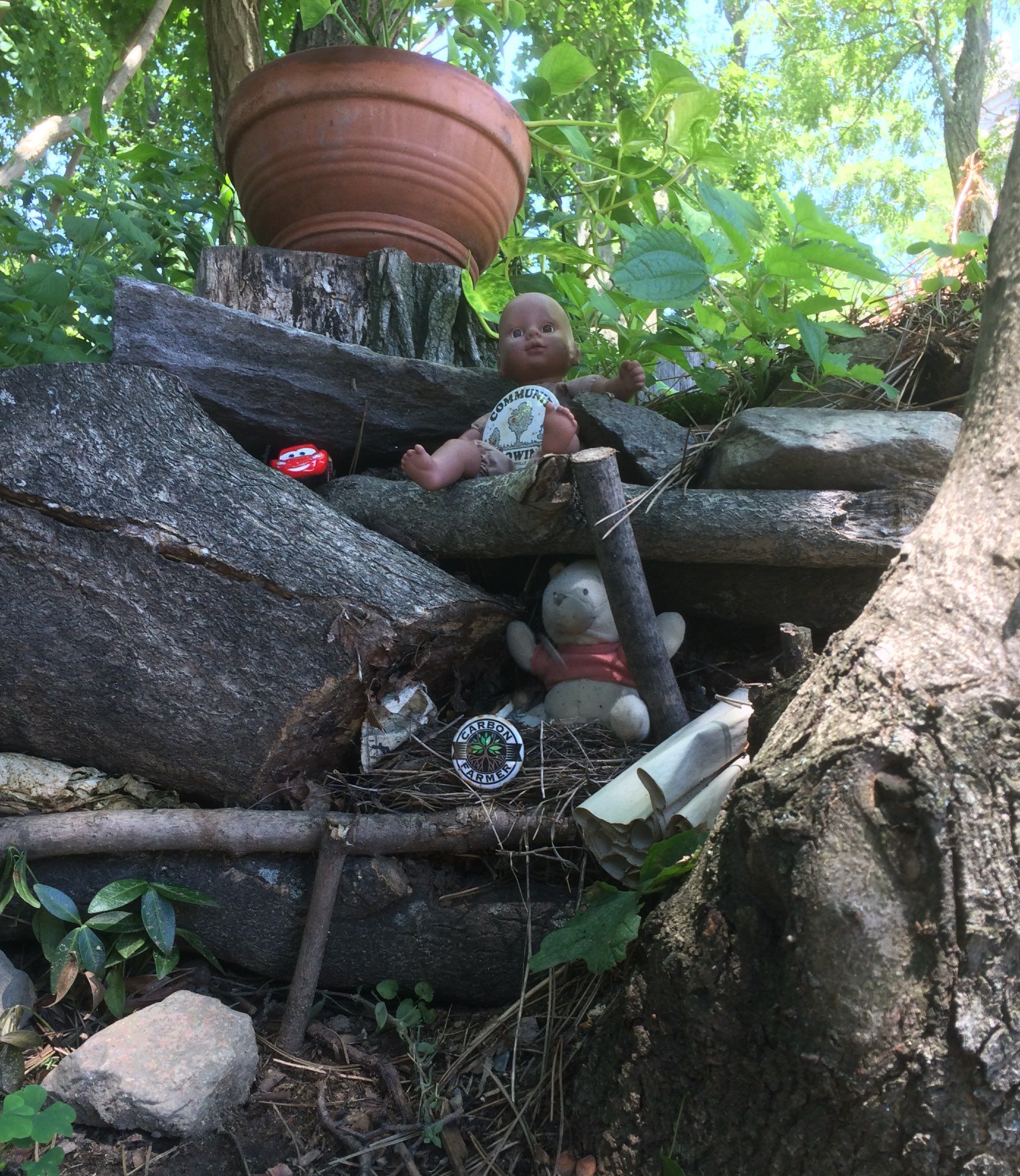 Close up on a nook formed by rocks and tree roots. A terra cotta plant pot sitting above the nook on a stump, then scattered below: a baby doll whose torso is covered with a badge that says "Community" and has a drawing of a tree, a red toy car with a face, as in the animated movie "Cars," a very faded little stuffed Winnie the Pooh, and a round badge that says "Carbon Farmer" around a center that has an image of green leaves in the middle.