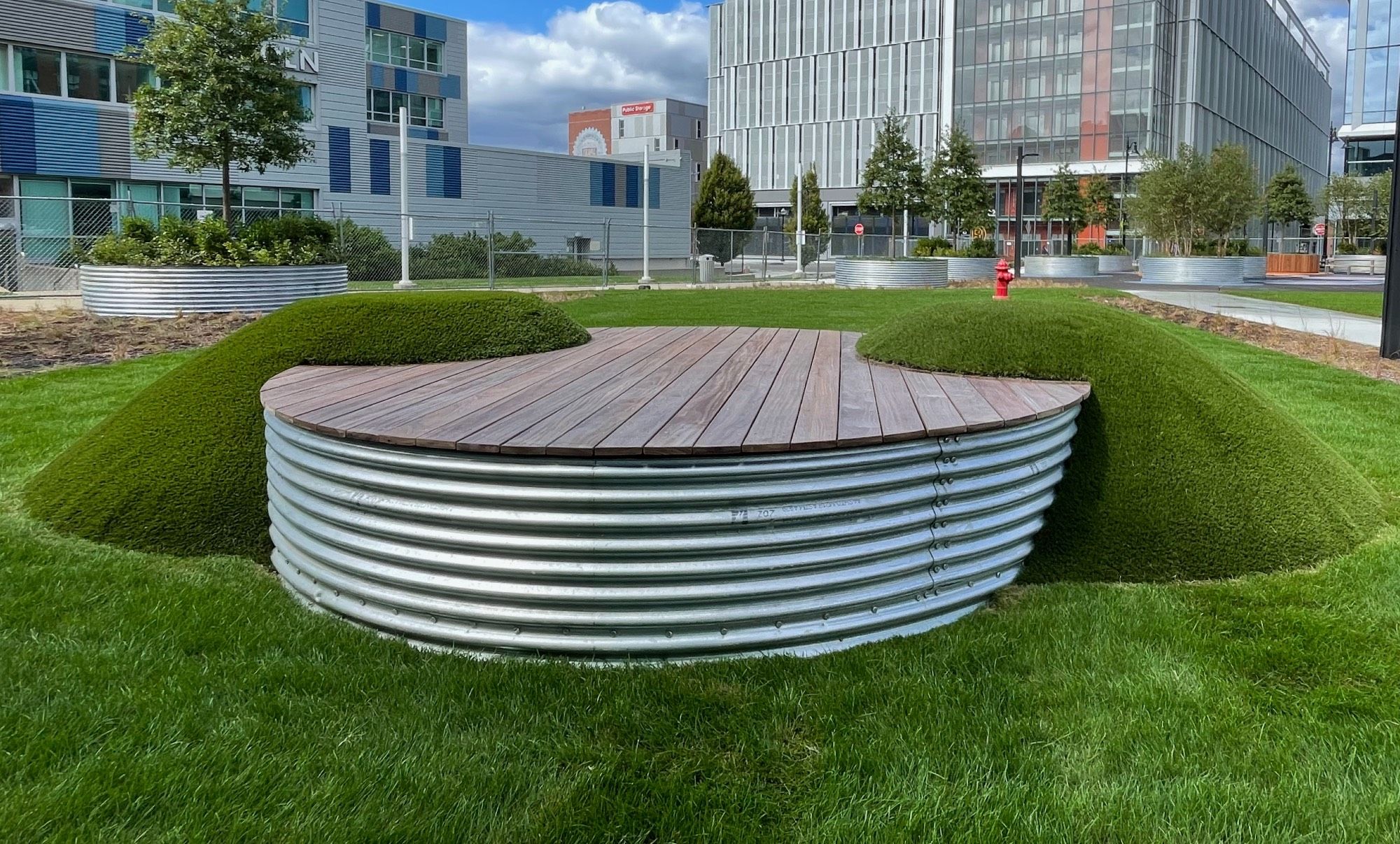 In a grassy are surrounded by large modern office buildings, we're looking at a round sort of platform maybe two feet tall, sides of corrugated aluminum all around, and flat top of planks of wood, like a deck, with surface areavthat could be occupied, say, by a 3-piece band. On either side a grass-covered hump intersects the platform, as if two grass creatures opened their mouths to take a bite out of this hockey-puck shaped thing.