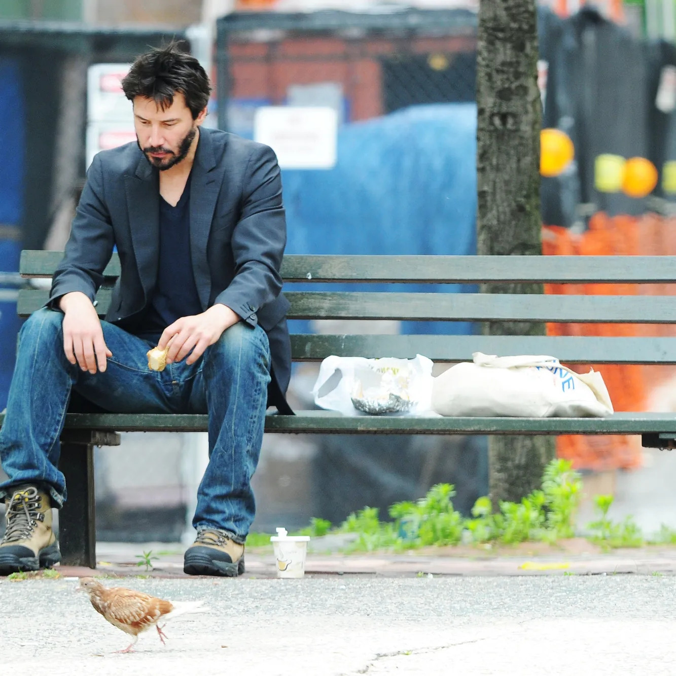 Sad Keanu Reeves sitting on park bench eating a sandwich while a scruffy bird ambles by hoping for crumbs