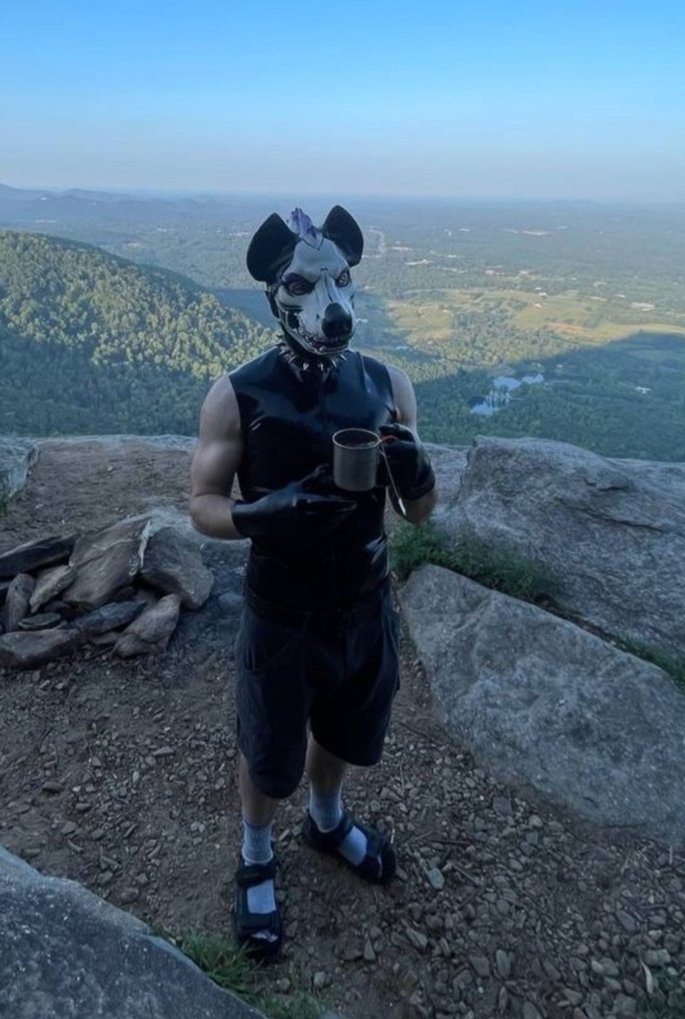 Irl photo of me, Skwuz, standing by a cliff and holding coffee with a sweeping vista of a valley behind me. I'm wearing a latex skull hyena hood and latex vest, spiked collar, hiking shorts, socks and sandals