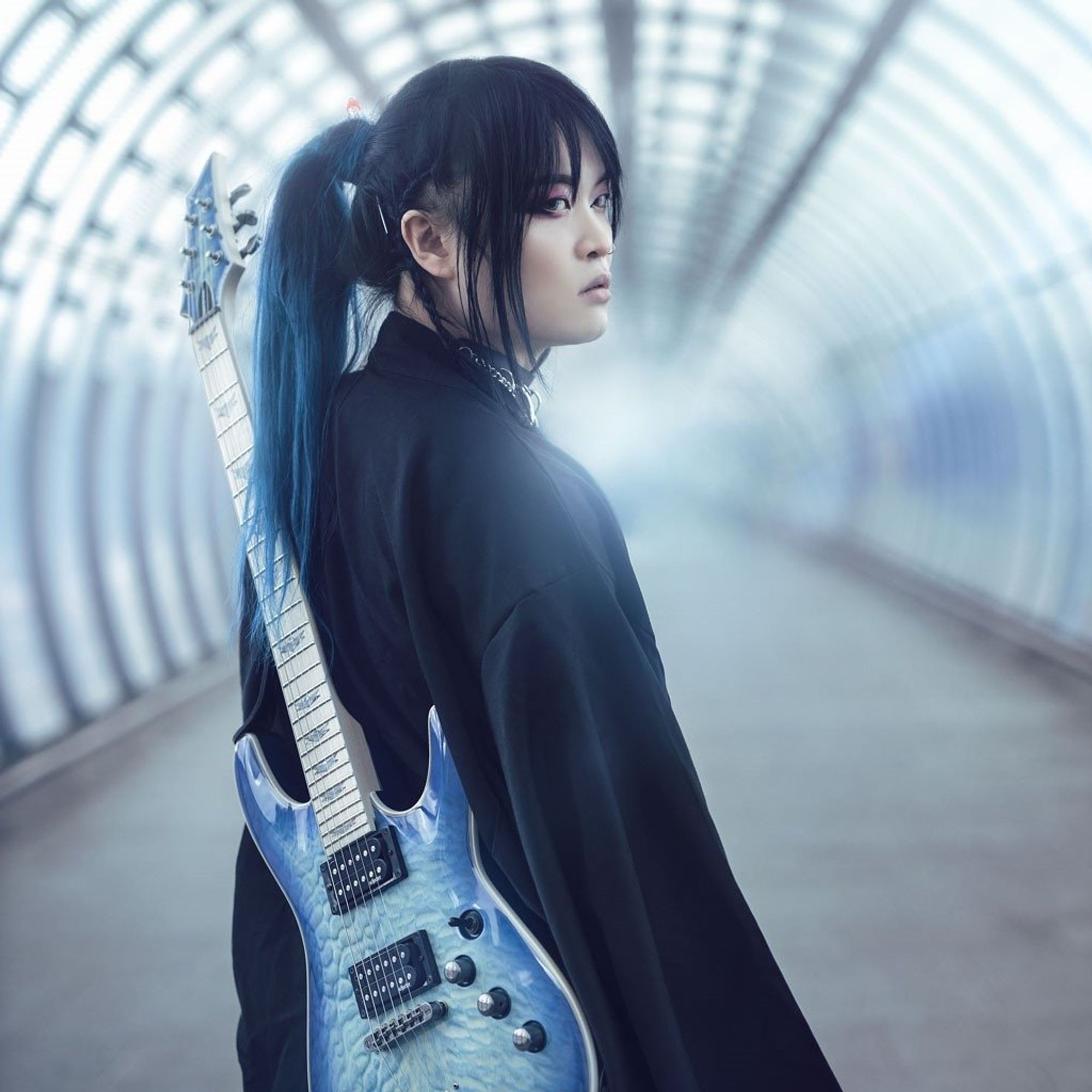 Band leader Kai is dressed in black clothing and carries a soft blue-coloured electric guitar on the back. Kai seems to be walking on an empty train station or in an empty tunnel and looks behind him.