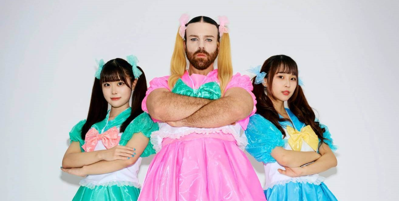 The three members of BABYBEARD, dressed in colourful dresses, in bright pink, blue and green are standing next to each other while having their arms crossed. LADYBEARD (in pink) stands in the centre of the photo and the two girls, Suzu (green) and Mahri (blue) are standing a bit behind him leaning on the heel of one of their shoes.