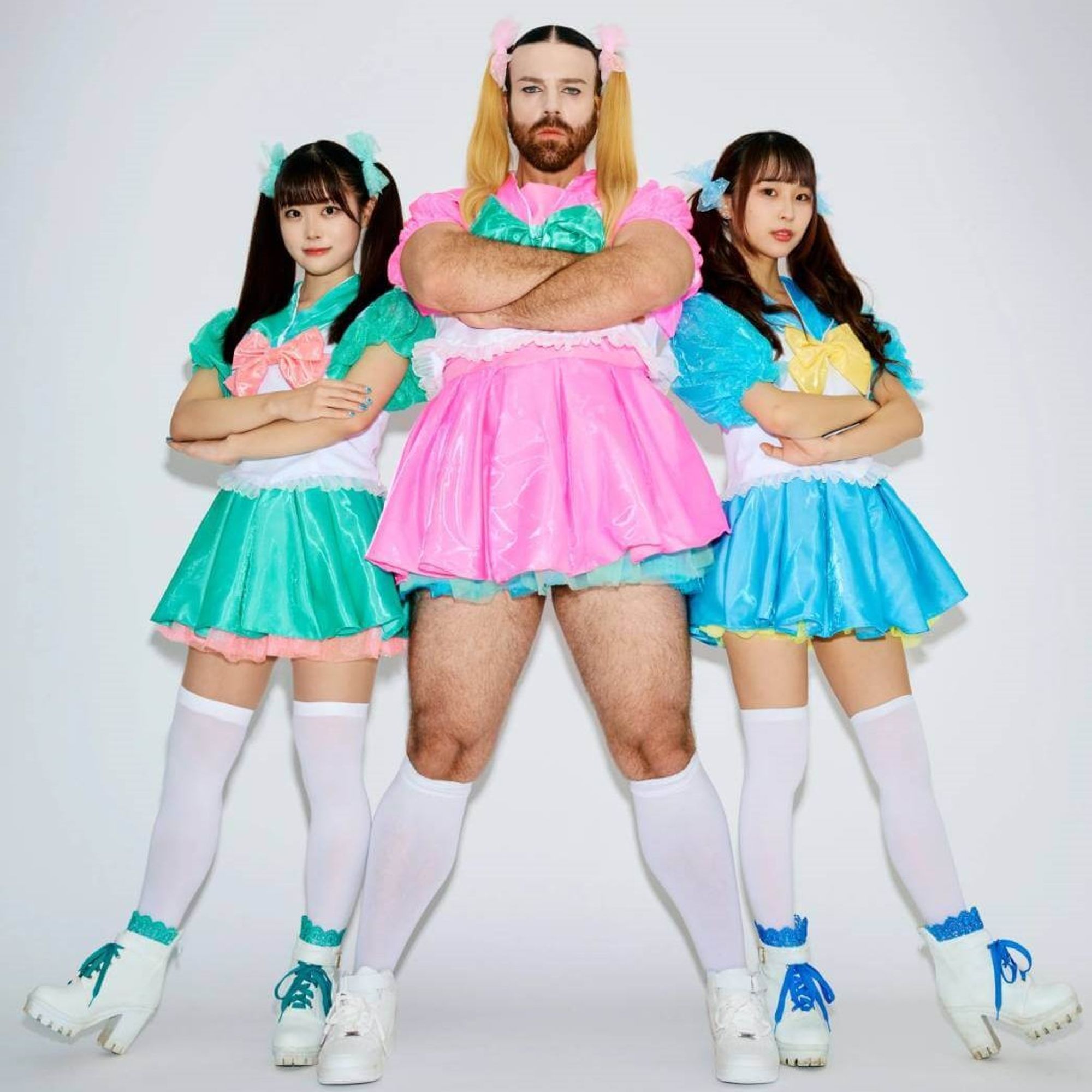 The three members of BABYBEARD, dressed in colourful dresses, in bright pink, blue and green are standing next to each other while having their arms crossed. LADYBEARD (in pink) stands in the centre of the photo and the two girls, Suzu (green) and Mahri (blue) are standing a bit behind him leaning on the heel of one of their shoes.