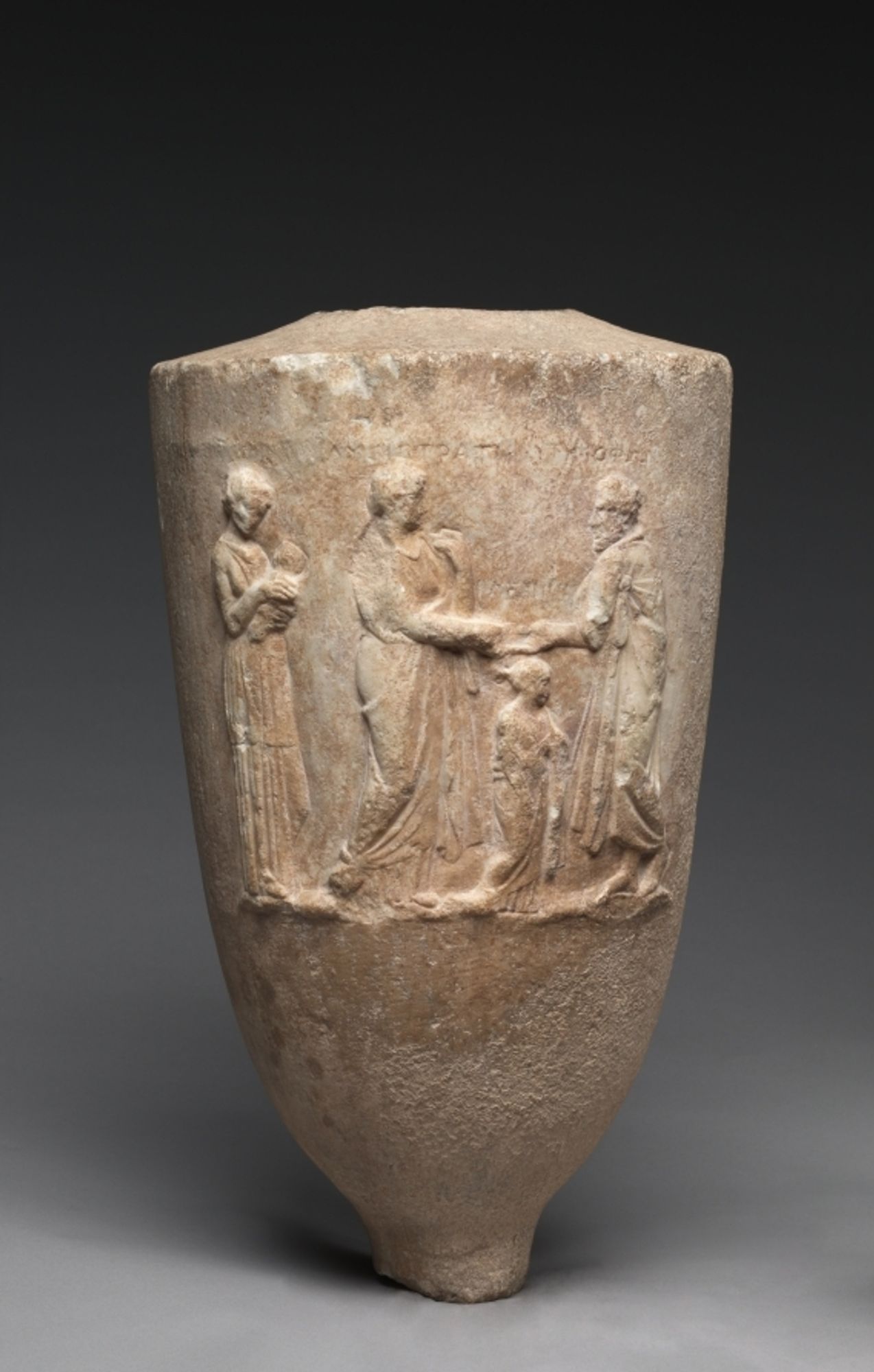 This Attic grave marker is in the form of a lekythos, a type of vase commonly left as a grave offering. On it is a scene in relief with inscriptions. At the right a bearded man wearing a himation (cloak), whose name “Timophon” is inscribed, clasps hands with a woman wearing a himation and mantle with the inscribed name “Lysistrate.” Below and between them is a girl identified by an inscription as “Kleippe.” At the left is an unidentified woman, likely a nurse, holding an infant. The scene probably commemorates Lysistrate’s death in childbirth. Her newborn child apparently survived.