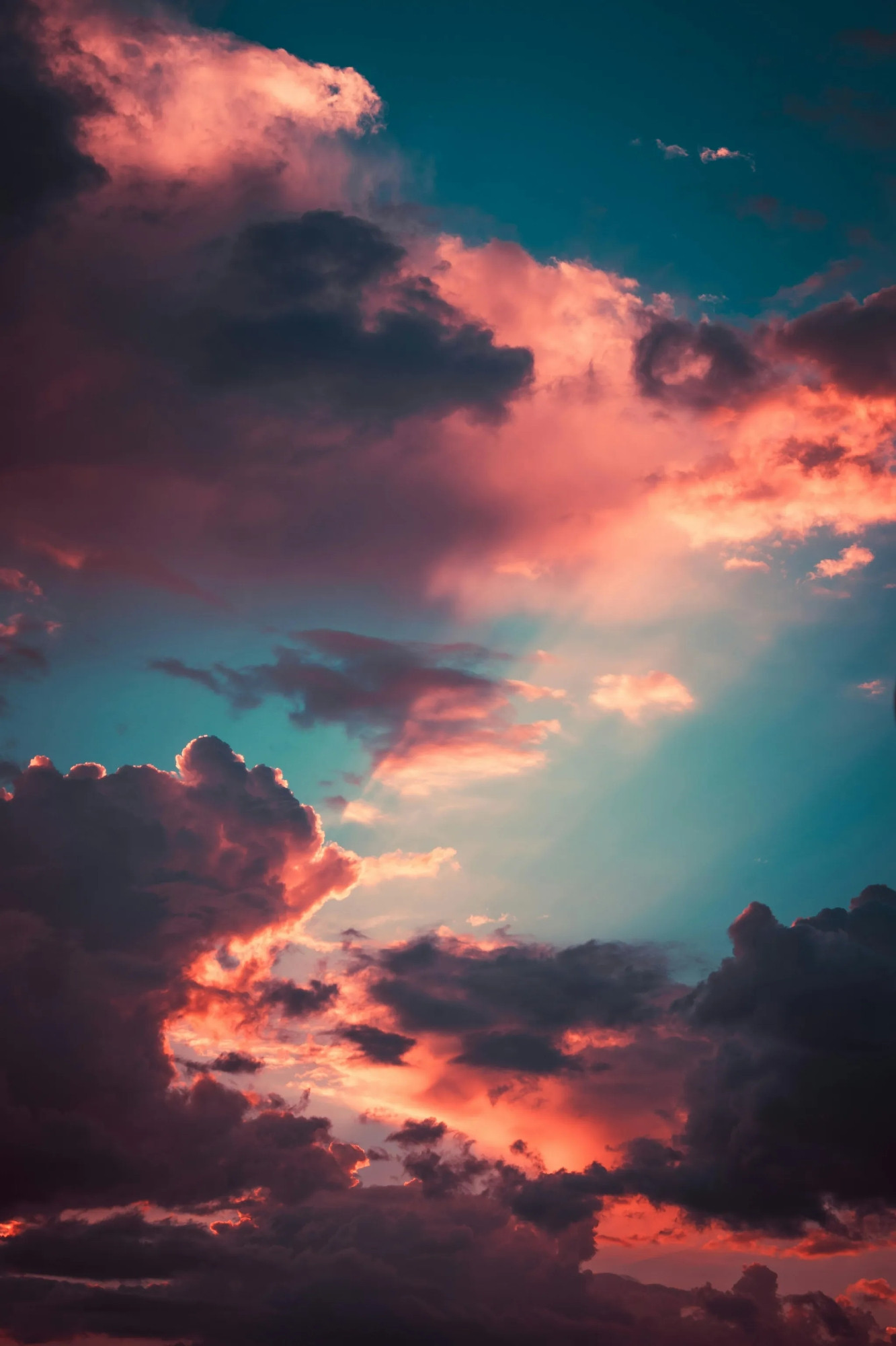 PHOTO by Szabó Viktor. The image showcases a dramatic sky at sunset, where deep blue and teal hues blend with warm shades of orange and pink. Billowing clouds, tinged with fiery highlights, create a striking contrast against the cooler tones of the sky. Sunlight filters through the gaps, casting a soft glow that enhances the depth and texture of the clouds, evoking a sense of tranquility and wonder.
