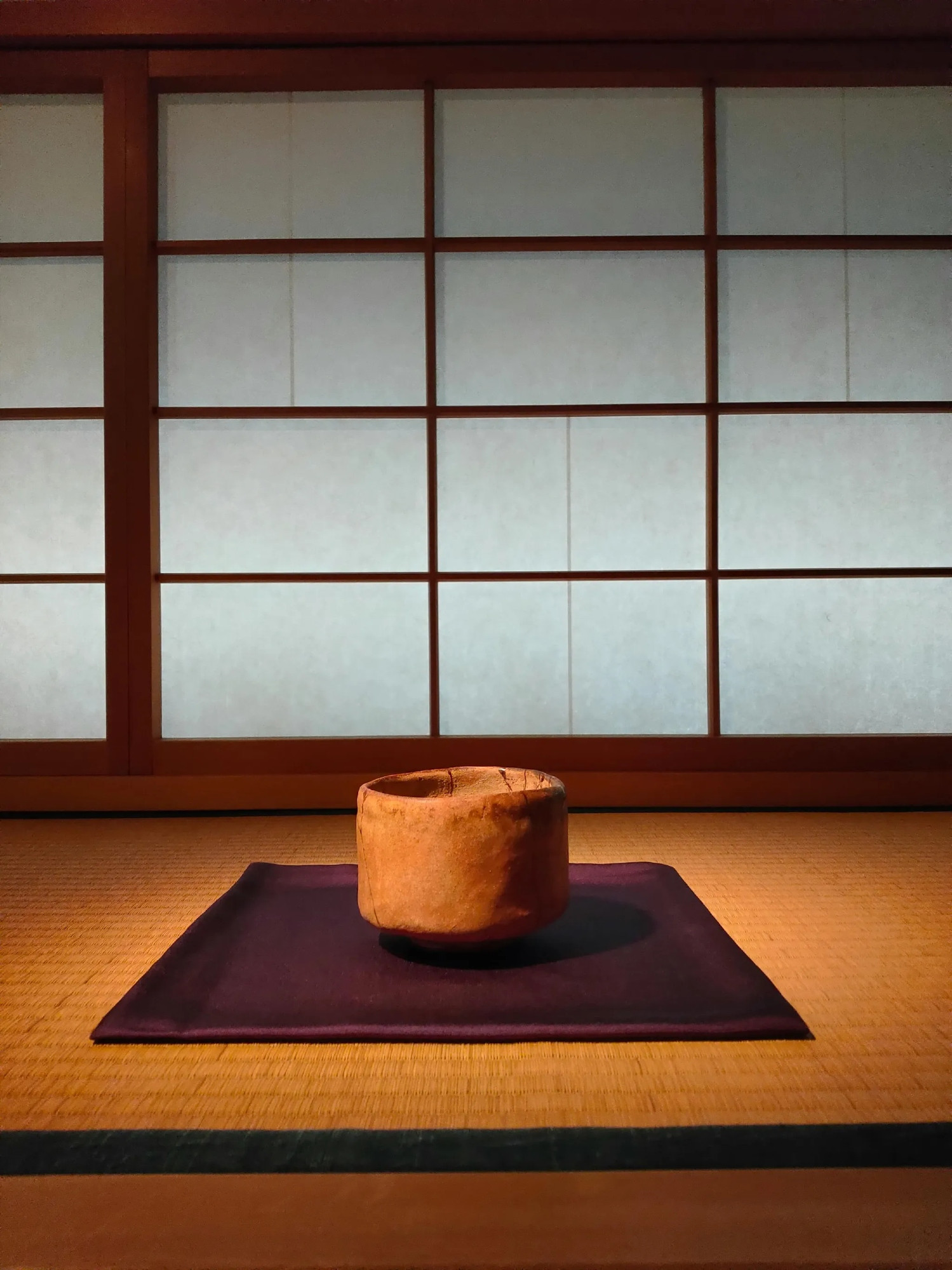 PHOTO by Daisuke Fujita. A rustic, earthy-toned tea bowl sits atop a deep purple cloth on a woven tatami mat, bathed in soft, natural light. Behind it, shoji screens filter a gentle glow, their wooden lattice framing a scene of quiet simplicity. The bowl’s imperfections and organic shape embody the spirit of wabi-sabi, inviting contemplation in the serene setting of a traditional Japanese tea room.