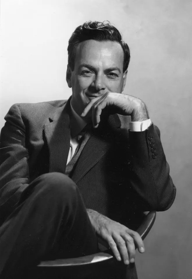 Renowned physicist Richard P. Feynman sits in a chair, one leg crossed, wearing a pinstripe suit. He rests his chin thoughtfully on his hand, eyes meeting the camera with a knowing smile. The black-and-white portrait captures a moment of relaxed brilliance, his posture casual yet alert, against a plain backdrop that highlights his presence.