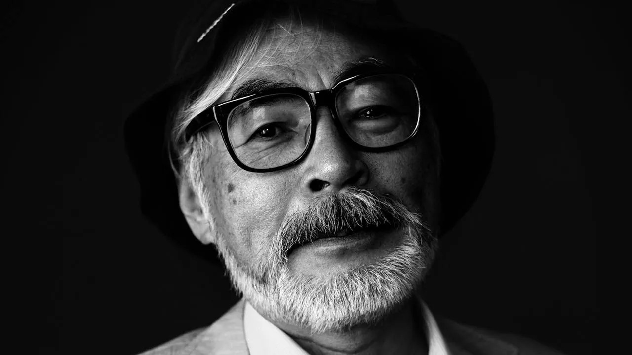 A black-and-white portrait of Hayao Miyazaki, the legendary animator and storyteller. His neatly trimmed white beard and thick-rimmed glasses frame a face full of wisdom and quiet determination. He wears a dark bucket hat, slightly tilted, and gazes with an introspective calm. The interplay of light and shadow enhances the depth of his expression, reflecting the mind behind worlds that have shaped the imagination of generations.