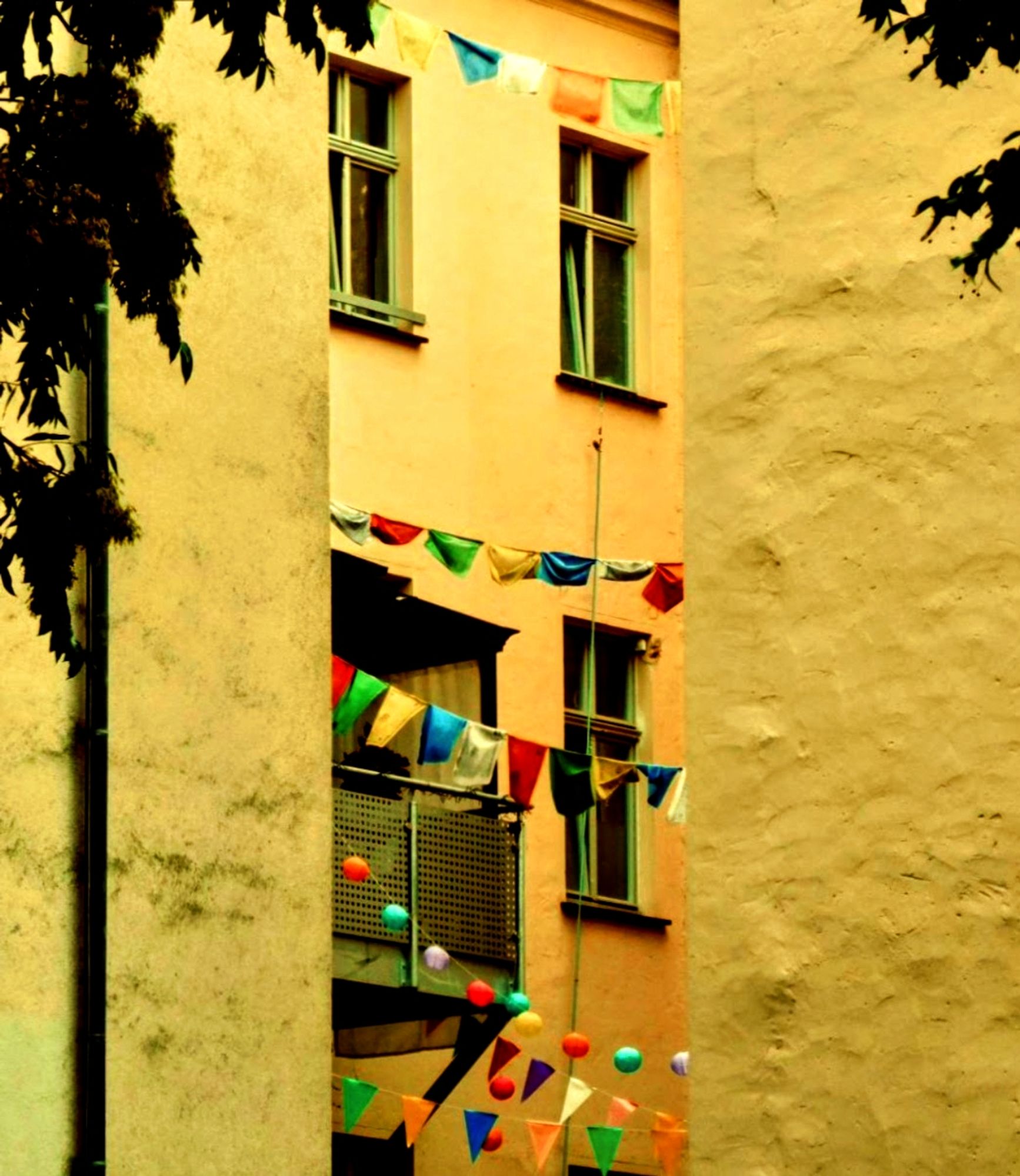 2 gelbe Hauswände , in deren Lücke nach hinten versetzt eine ebenfalls gelbe Hauswand zu sehen ist.Die Fenster haben grüne Fensterrahmen.Bunte Wimpelketten sind im Hinterhof zu sehen.