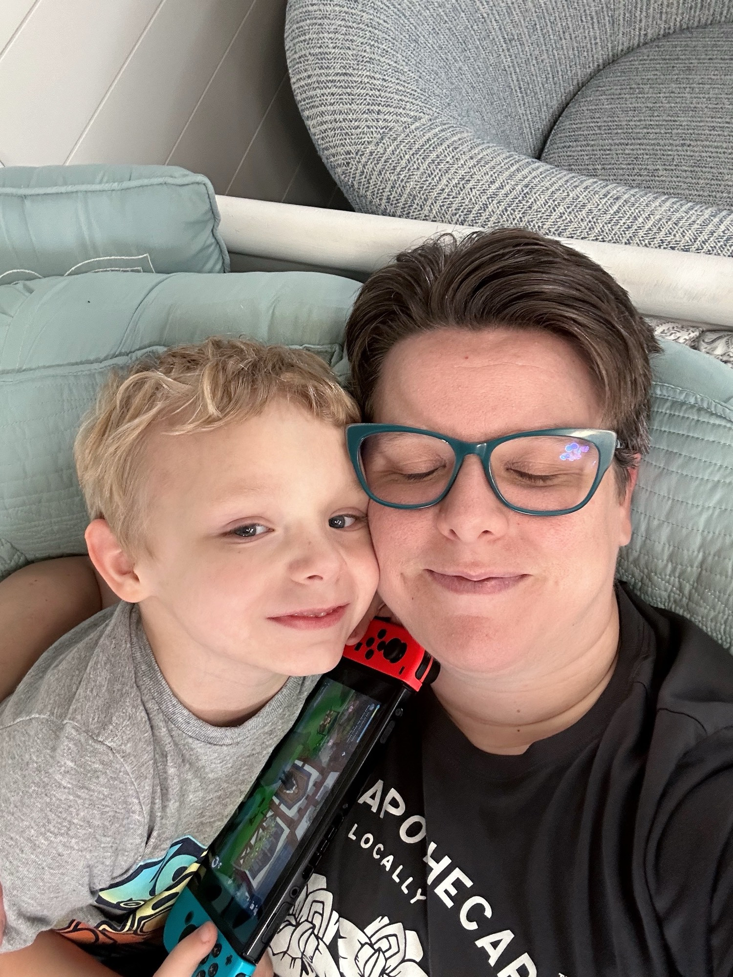 Eilonwy & kid chilling on day bed. Both are fair skinned. Eilonwy’s eyes are closed happily behind green glasses. Their kid smiles clutching a Nintendo switch. If you’ve watched Schitt’s Creek you might recognize my Rose Apothecary t shirt.